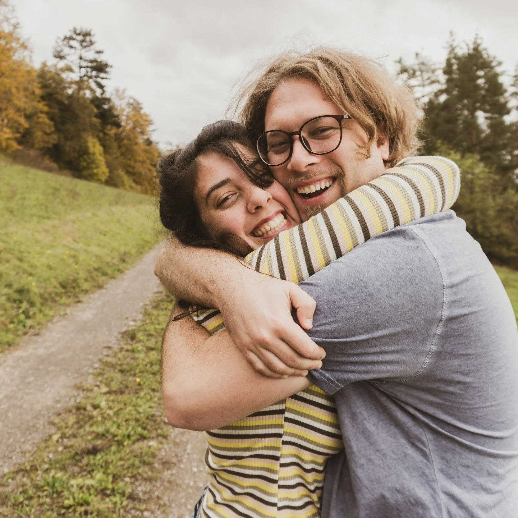 A happy couple hugging outdoors on a trail with trees and overcast sky in the background.