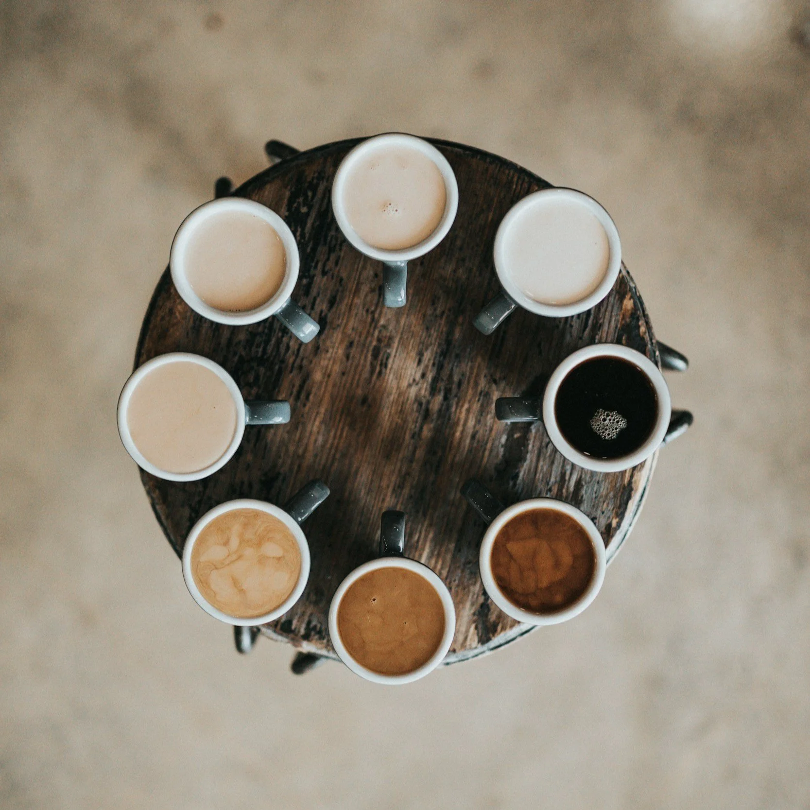Overhead view of a wooden table with nine ceramic mugs, each filled with different shades of coffee, arranged in a circle.
