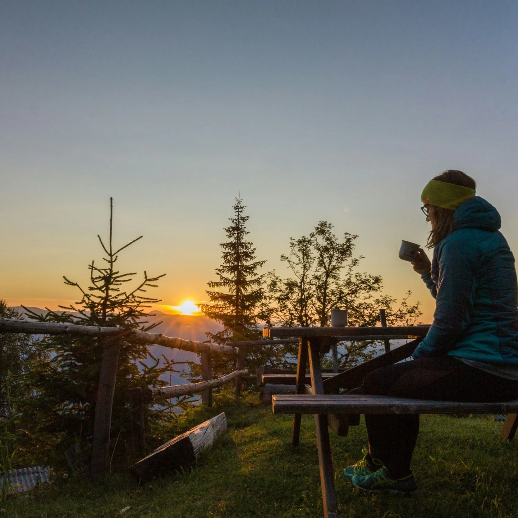 Person sitting at picnic table, drinking from a cup, watching sunrise or sunset over trees and mountains.