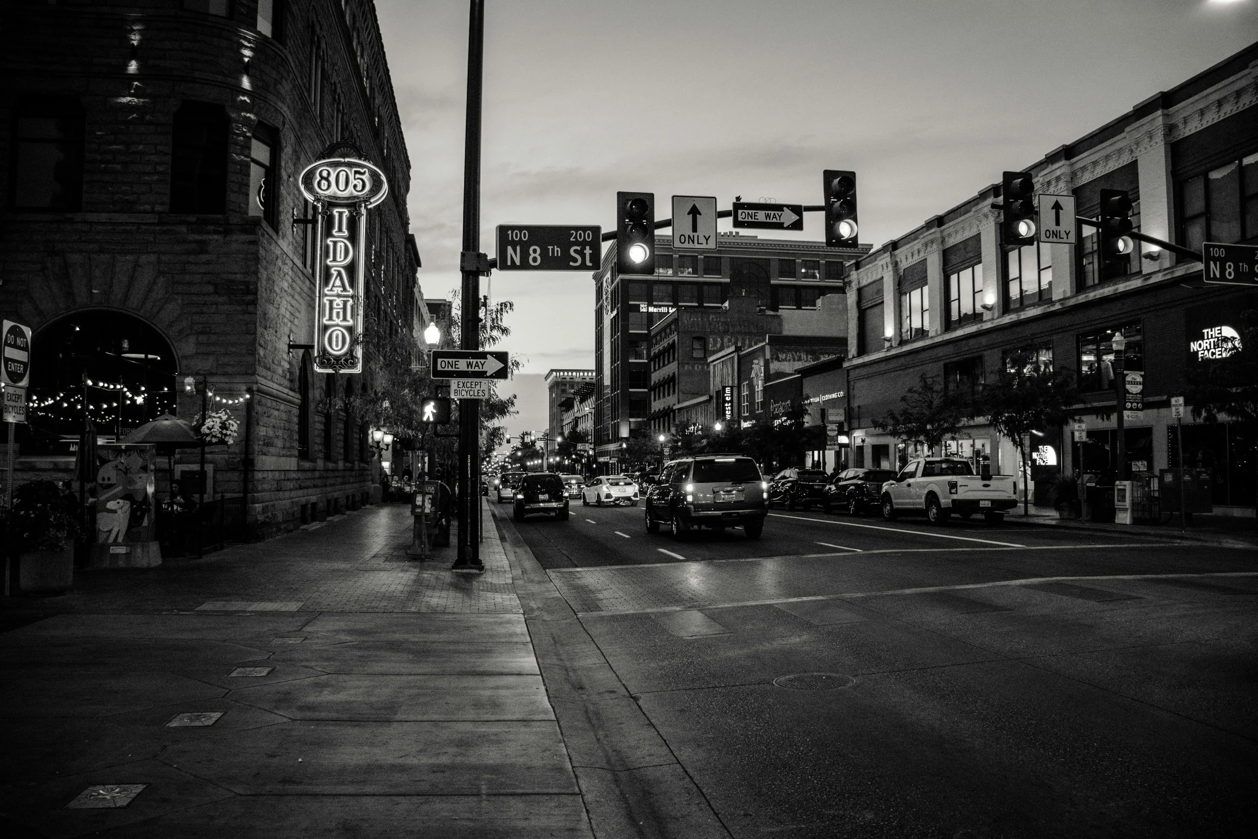 Downtown city street at dusk with cars, traffic lights, and buildings, including a neon sign for Idaho, in black and white.