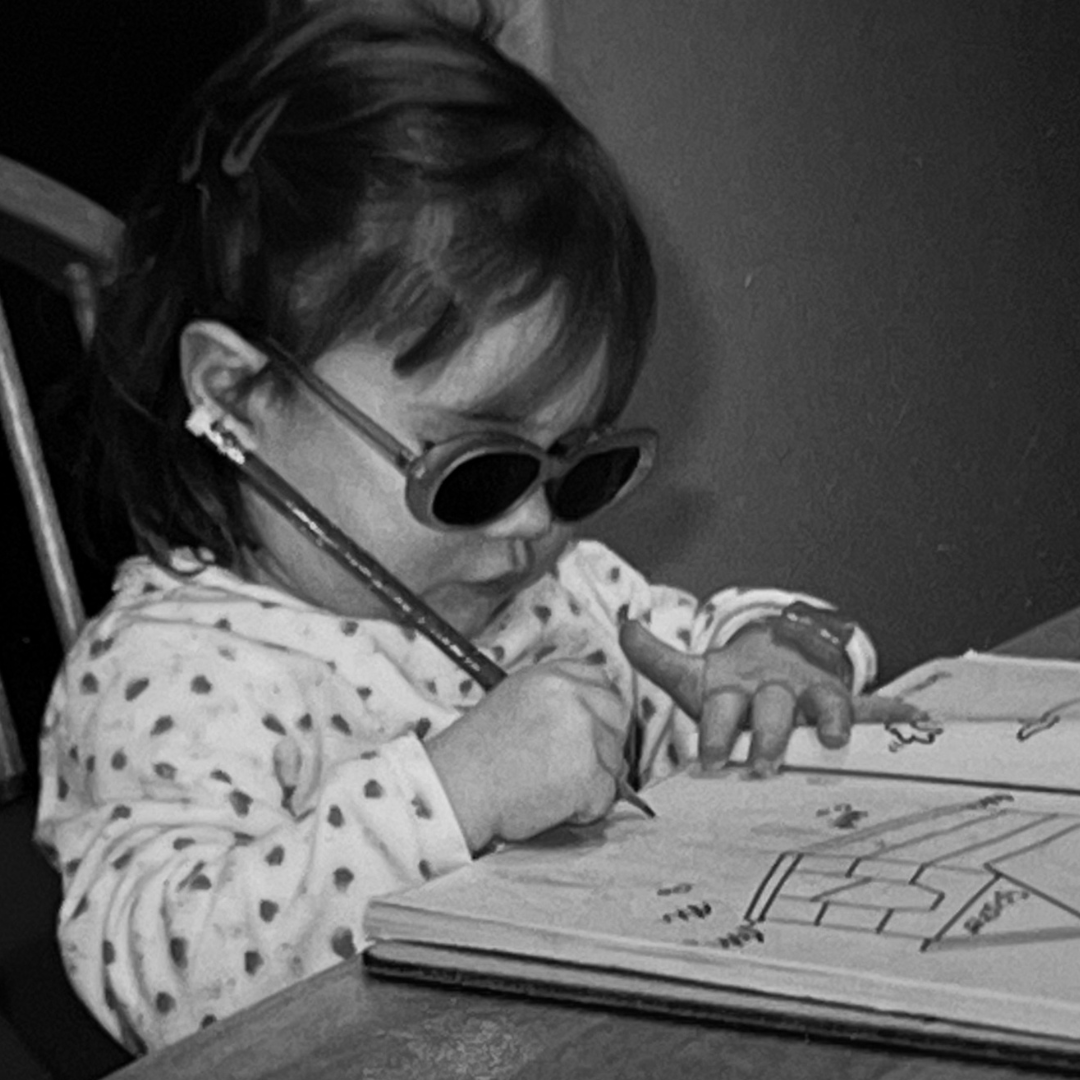 A young girl with short hair, wearing sunglasses and earrings, is focused on drawing and coloring in her book. She is sitting at a table.