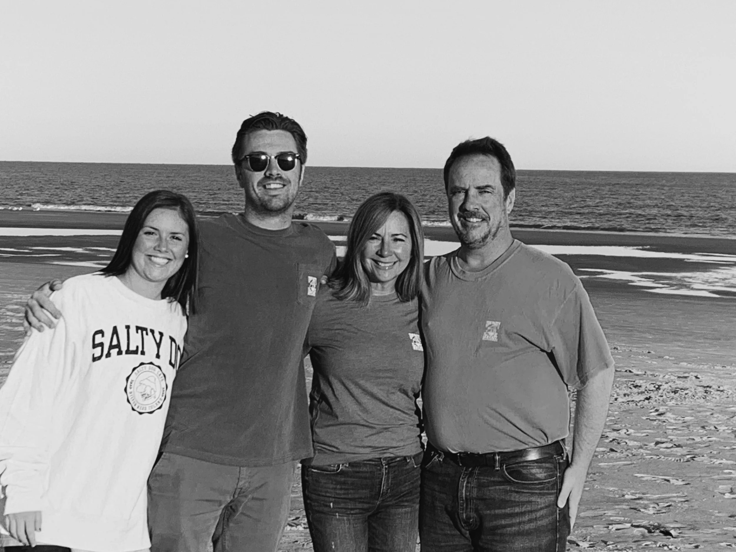 A group of four people standing together on a beach, smiling. The background shows the ocean and the sandy shoreline.