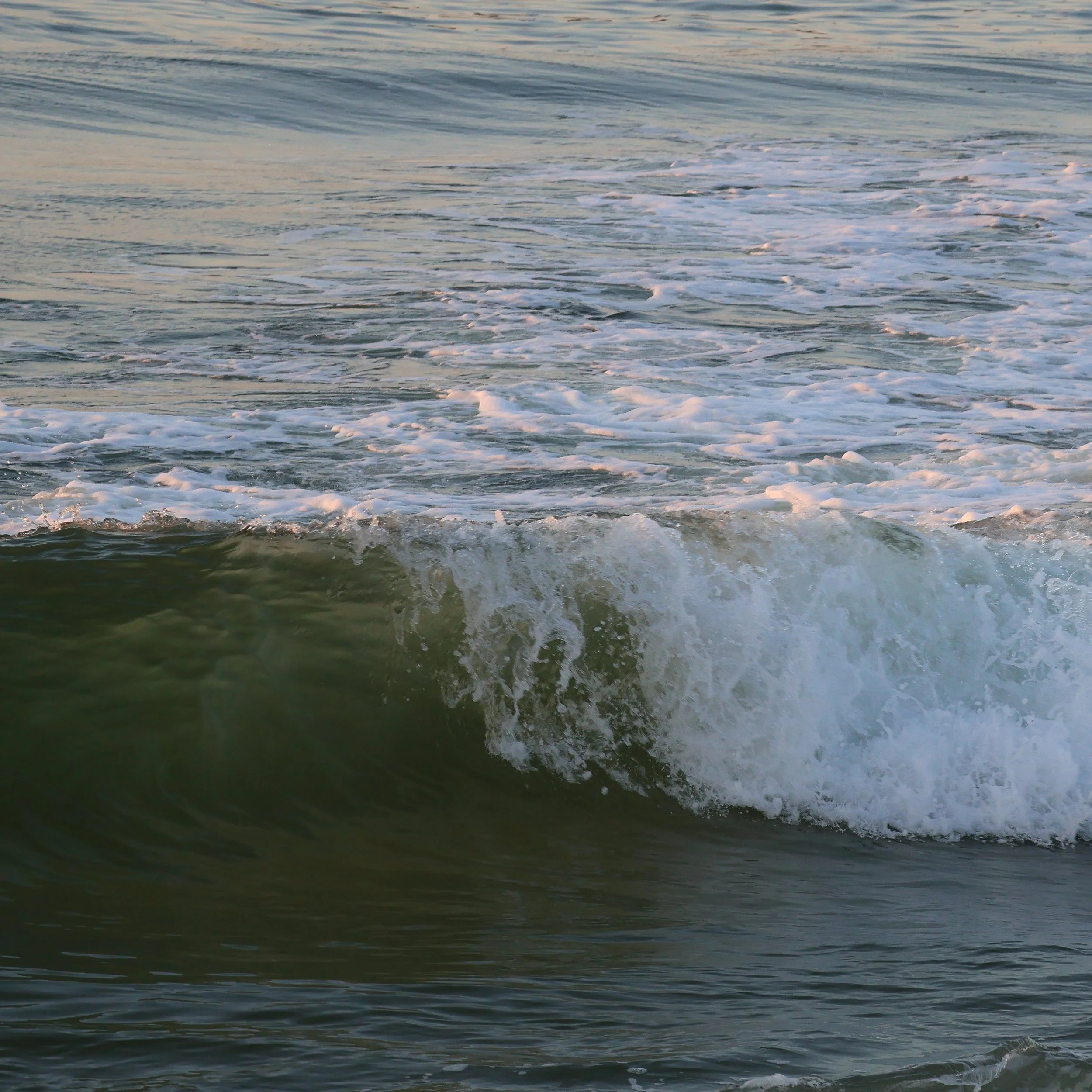 A photo of ocean waves with white foam and ripples on the water surface during sunrise.