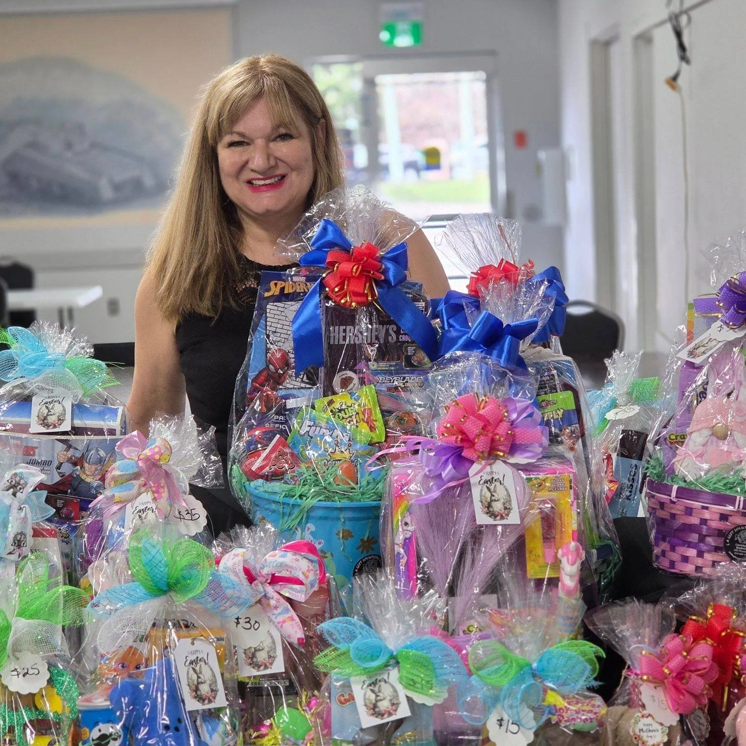 A smiling woman with long light brown hair standing behind a table filled with colorful wrapped Easter baskets, decorated with ribbons and bows, in a room with a door and a painting on the wall.