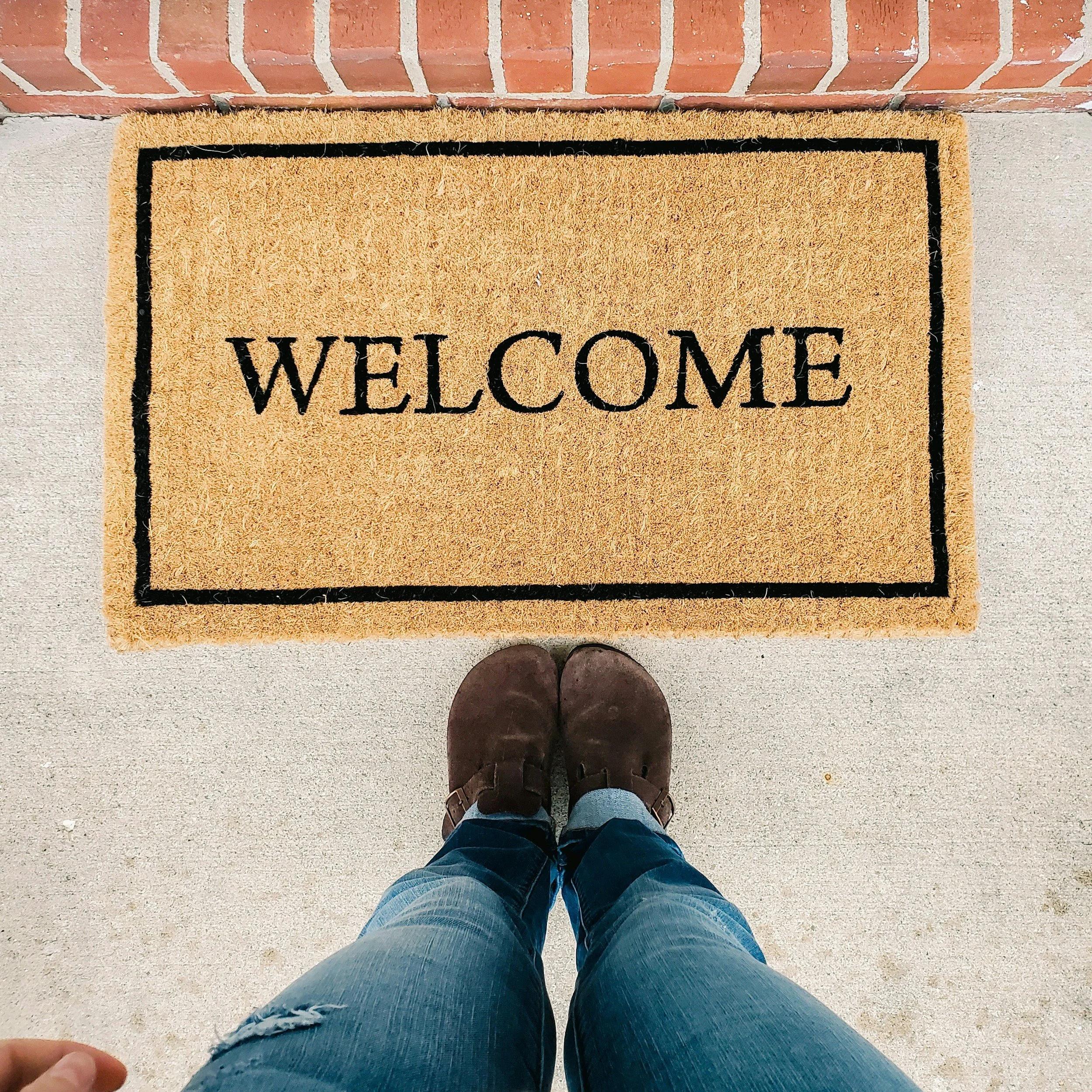 Top view of a person standing on a front porch, wearing blue jeans and brown shoes, looking down at a welcome mat and a concrete sidewalk.