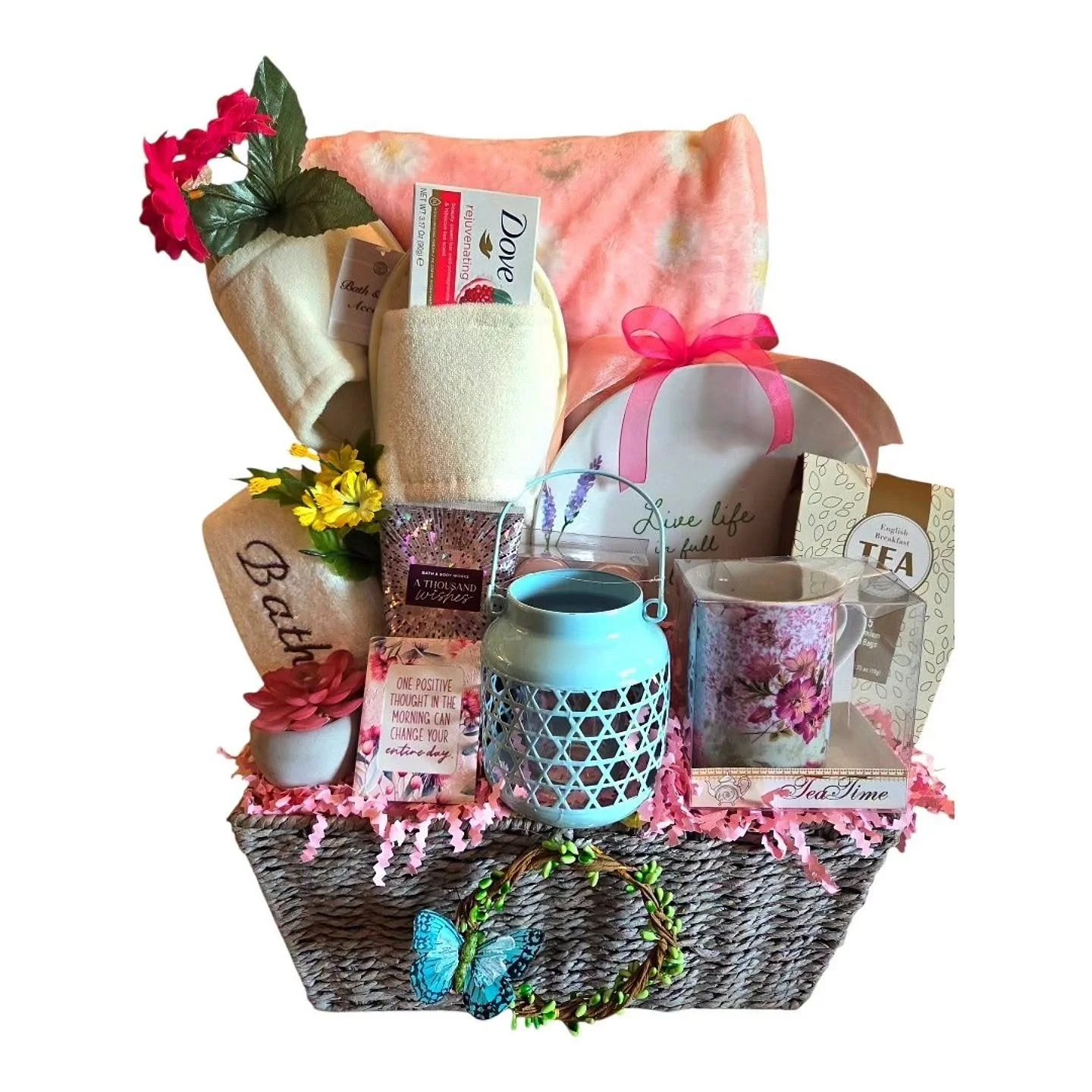 A gift basket filled with a pink blanket, a white towel, a box of Dove rejuvenating soap, a small bouquet of yellow flowers, a white lantern, a mug with floral design, a box of English breakfast tea, a pink flower-shaped candle, and various decorative items, all arranged on a pink paper bedding.