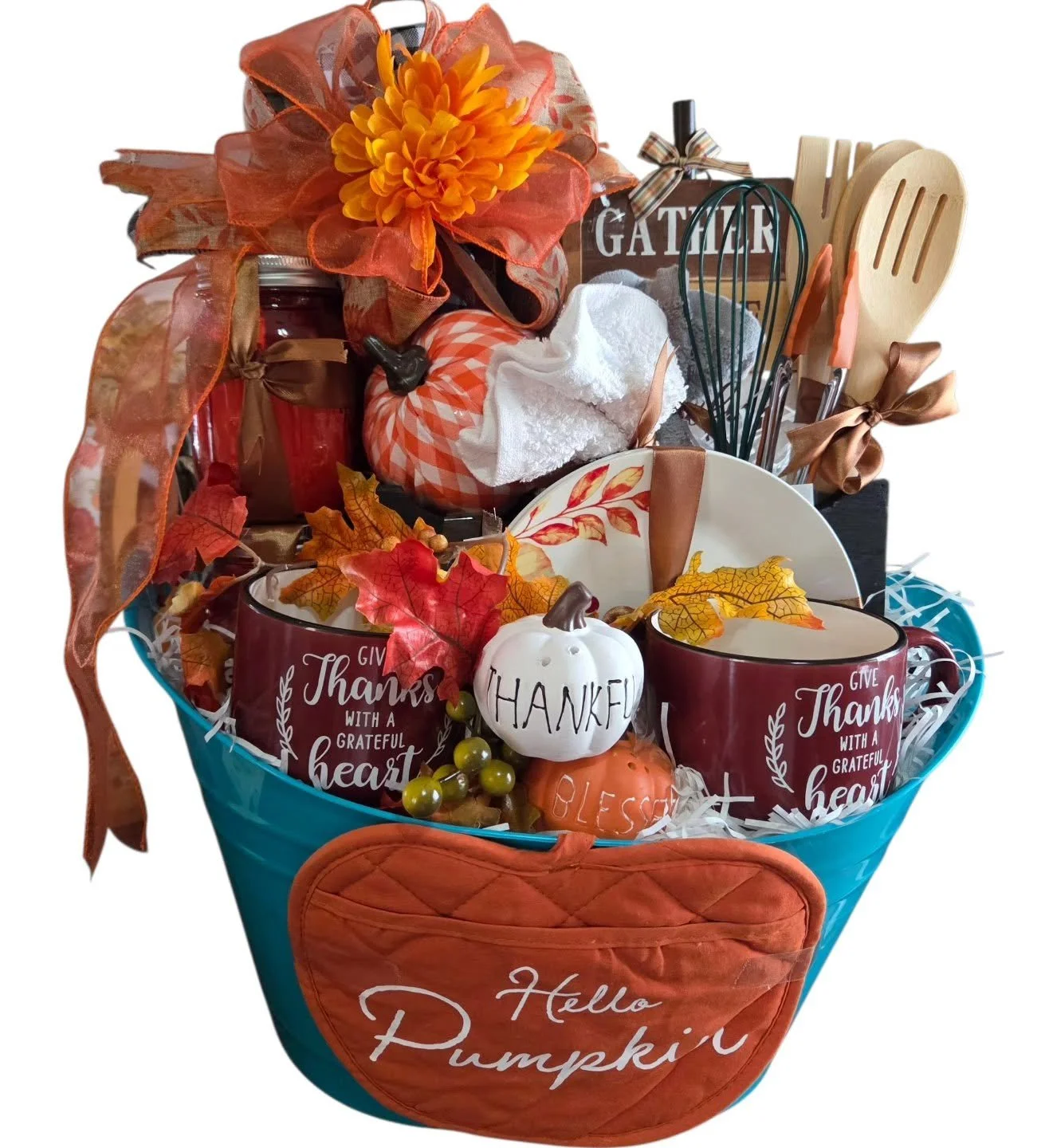 A welcome pumpkin-themed gift basket with autumn decorations, including a white pumpkin, fall leaves, mugs with gratitude messages, kitchen utensils, a jar, a tissue roll, and artificial fall leaves, all arranged in a teal basket with an orange quilted pocket.