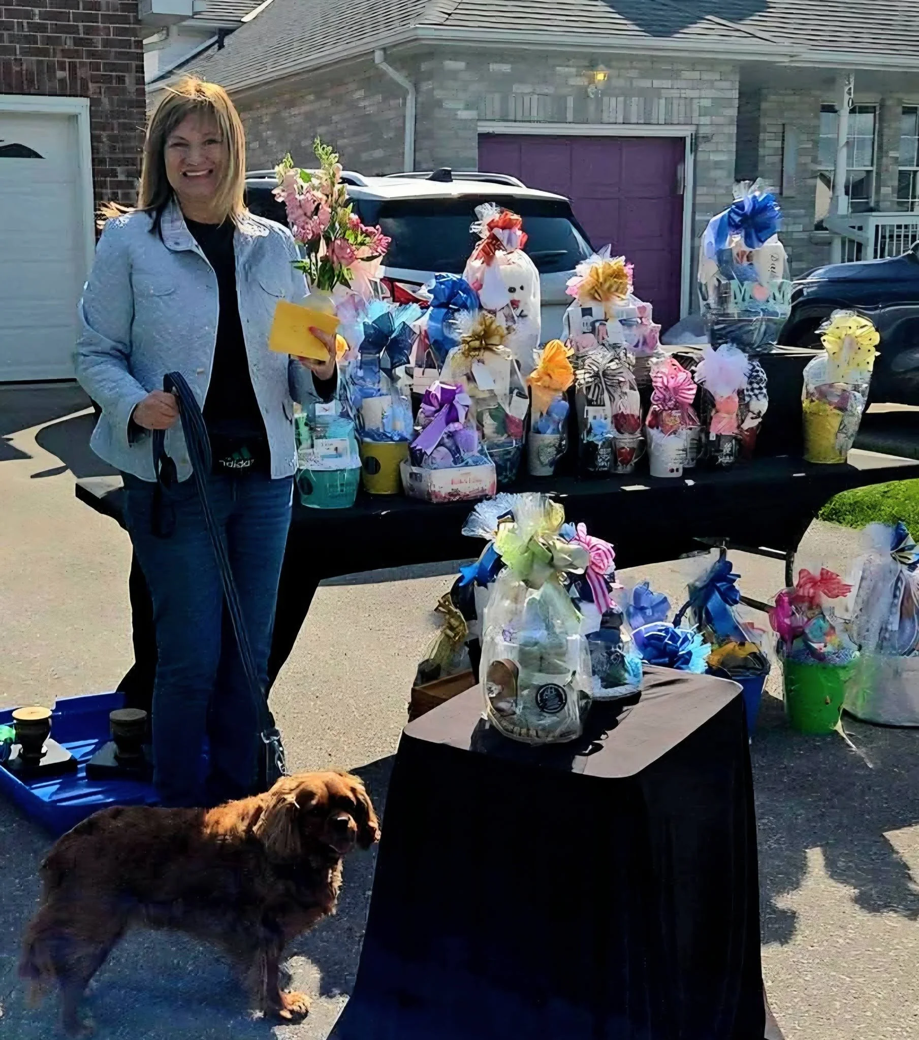 Woman standing outdoors near a table with wrapped gifts and a dog, with a parked car and house in the background.