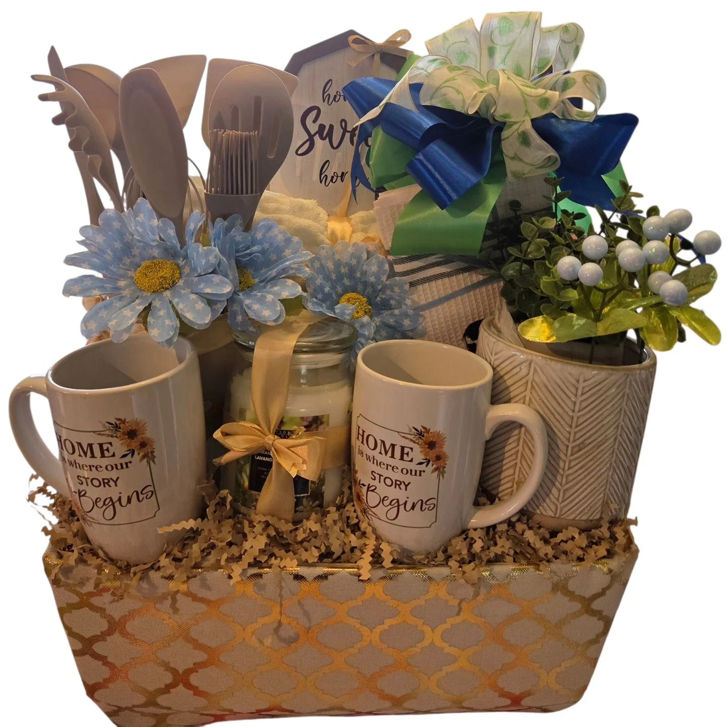 Decorative gift set including two white mugs with a floral quote, a jar with a scented candle, artificial flowers, and a wrapped gift box with a bow, all arranged on a patterned tablecloth with shredded paper filler.