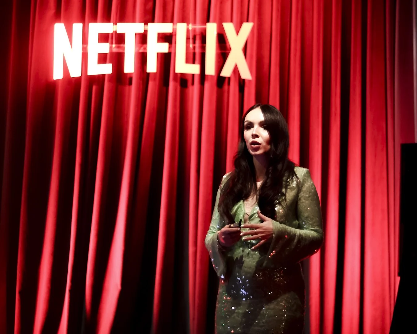 A woman with long dark hair wearing a green sequined dress standing on stage in front of red curtains and a large bright 'Netflix' sign.