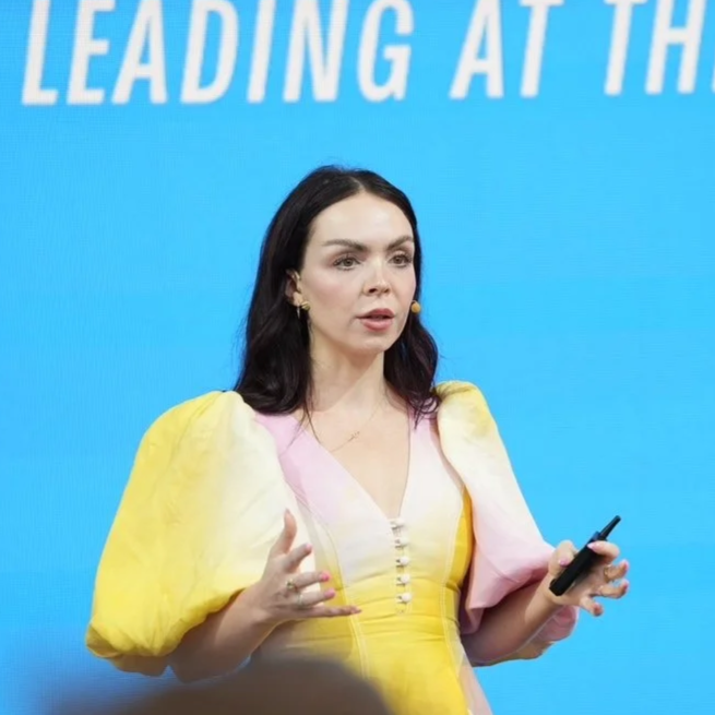 A woman with dark hair speaking at an event, holding a remote control in her right hand, against a blue background with text partially visible.