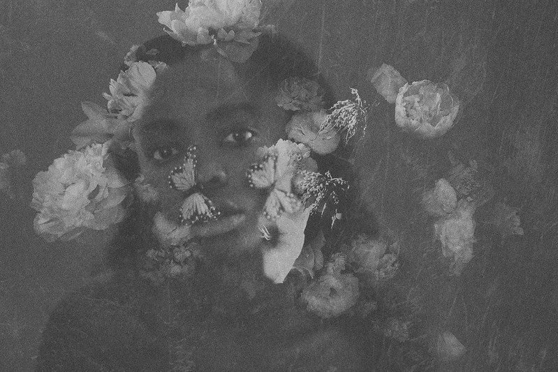 A black and white photo of a woman with flowers around her head and face, with butterflies on her face and in front of her eyes, creating a surreal, artistic composition.