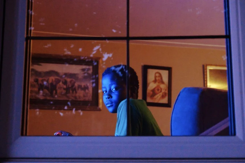 A young boy with braided hair looking out the window at night, illuminated by blue and orange lighting inside. There are framed pictures on the wall behind him.