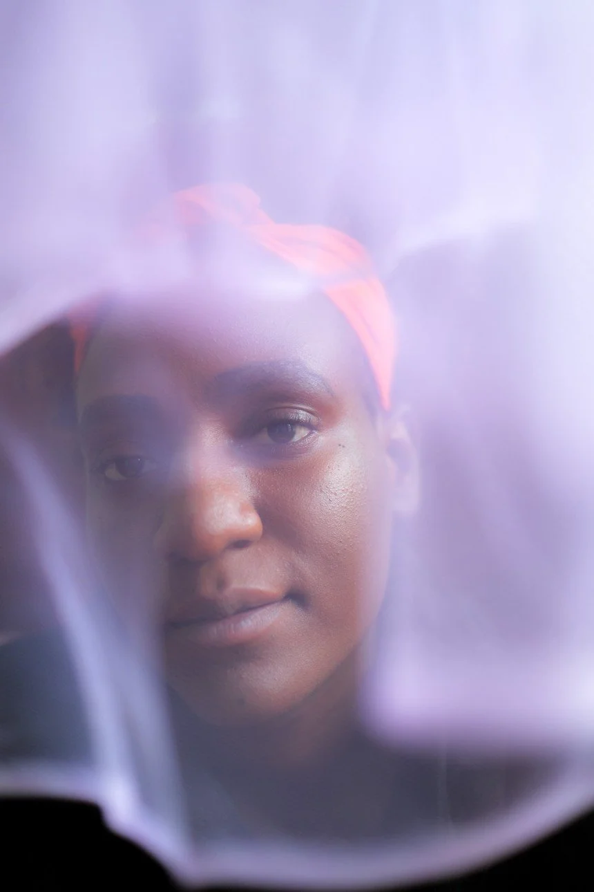 Close-up of a woman with dark skin and a colorful headscarf, partially obscured by semi-transparent fabric or material.