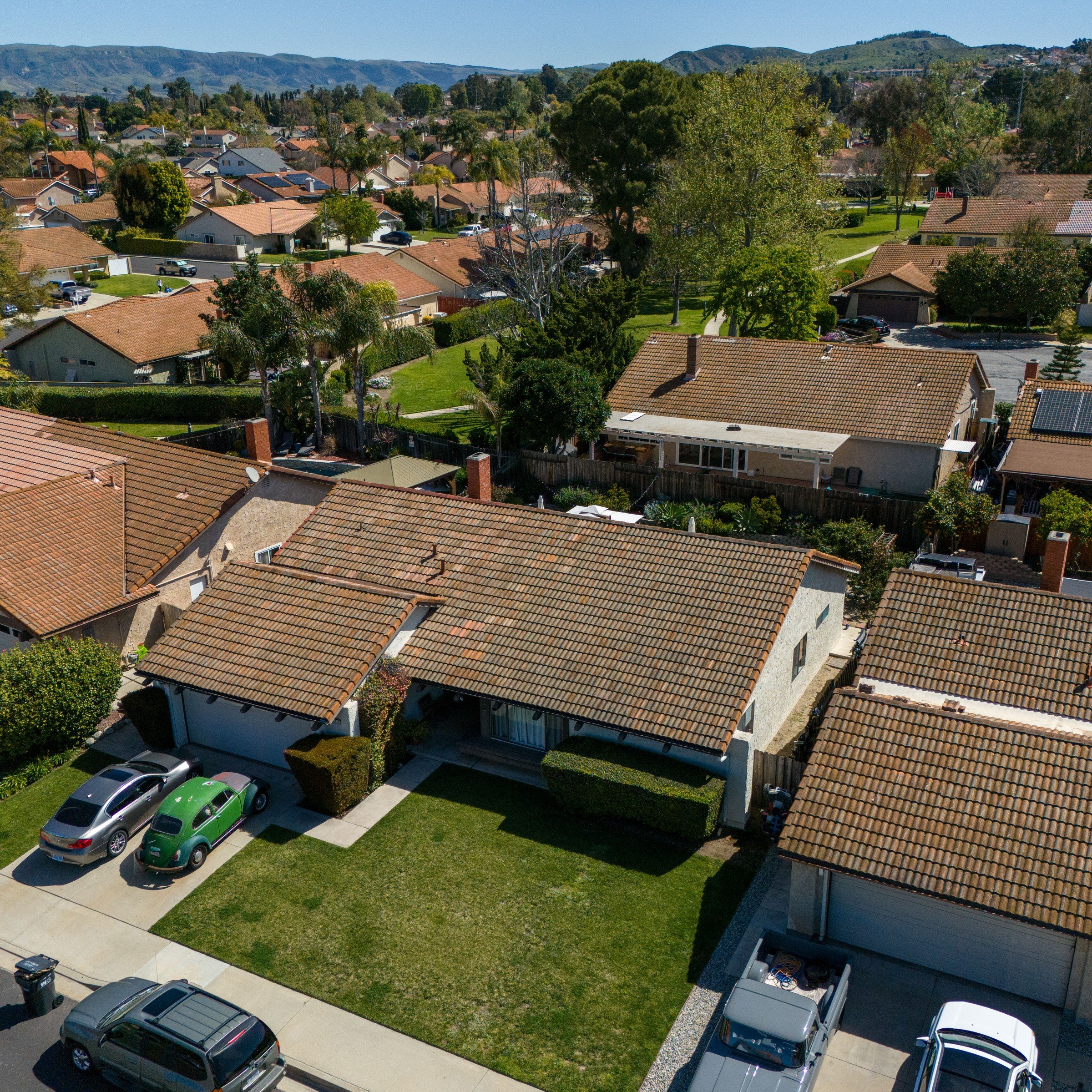 Aerial of a Moorpark neighborhood with brown tile roofs, manicured lawns, and hillside backdrop under clear sky