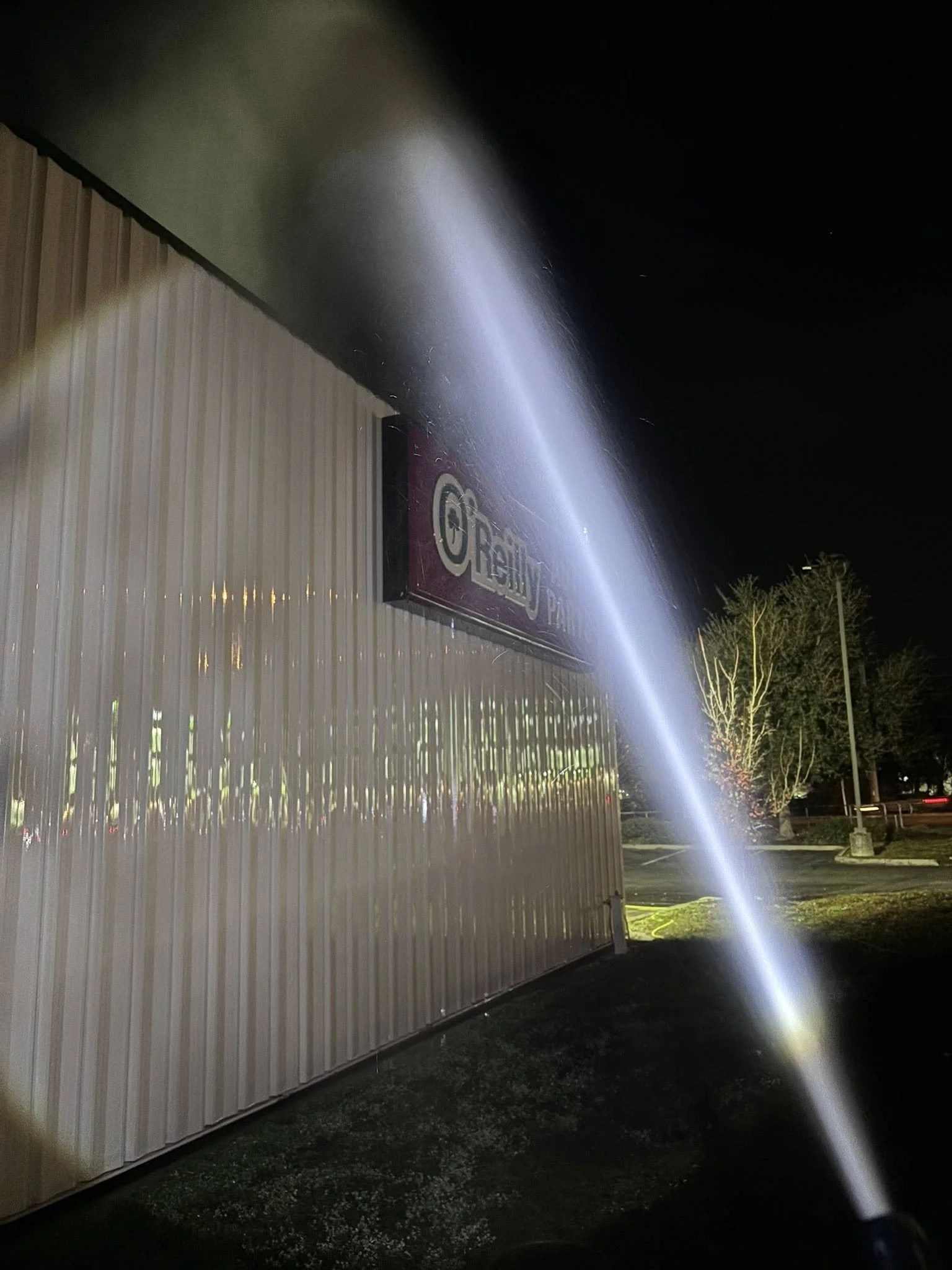 A night scene showing a beam of light from a flashlight hitting the side of a metal building with a sign that says "Beauty" and part of the parking lot with trees in the background.