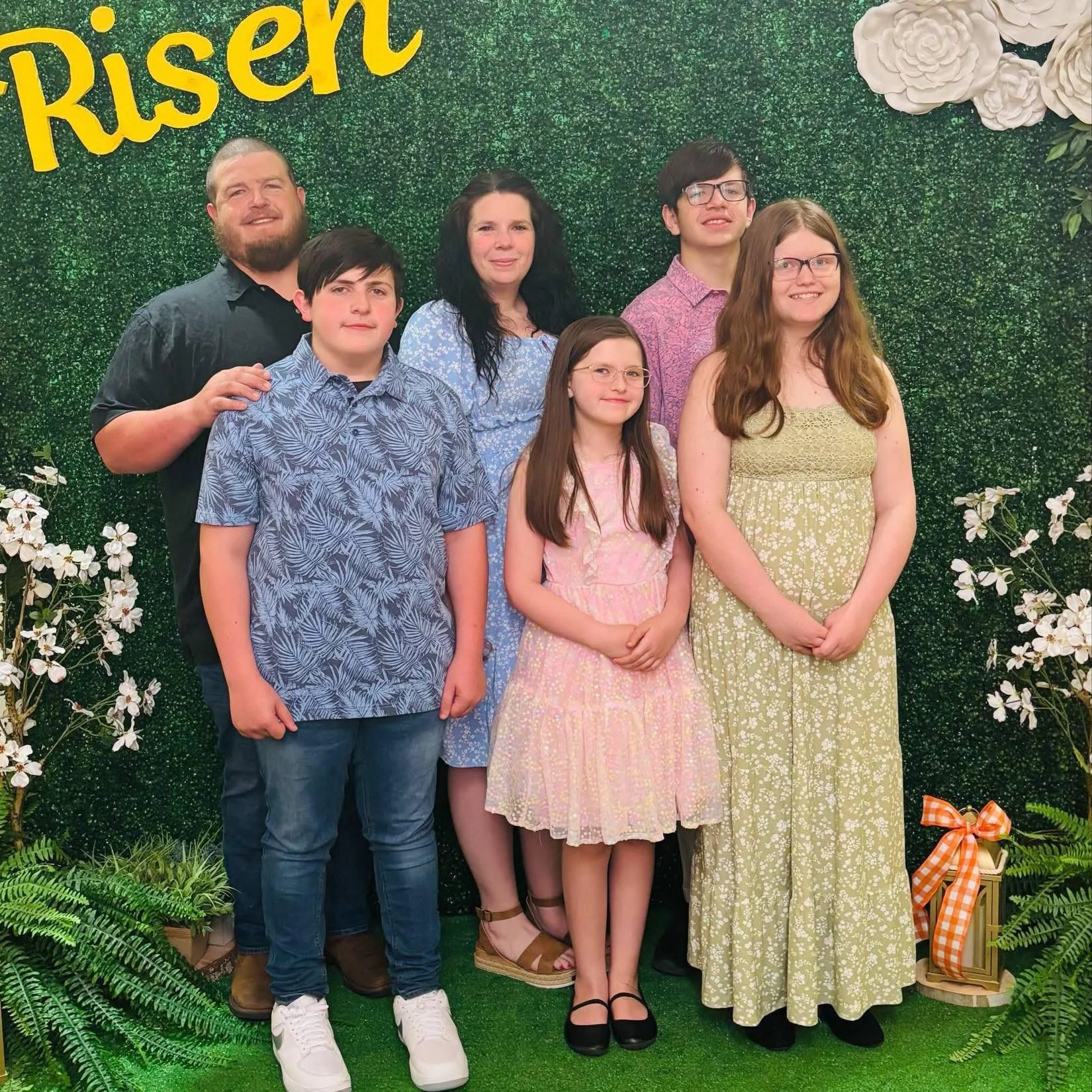 A family of seven posing in front of a decorated green backdrop with the word 'Risen' in yellow at the top left corner. The background features white flowers and greenery. The family members are dressed in casual summer clothing, smiling for the photo.