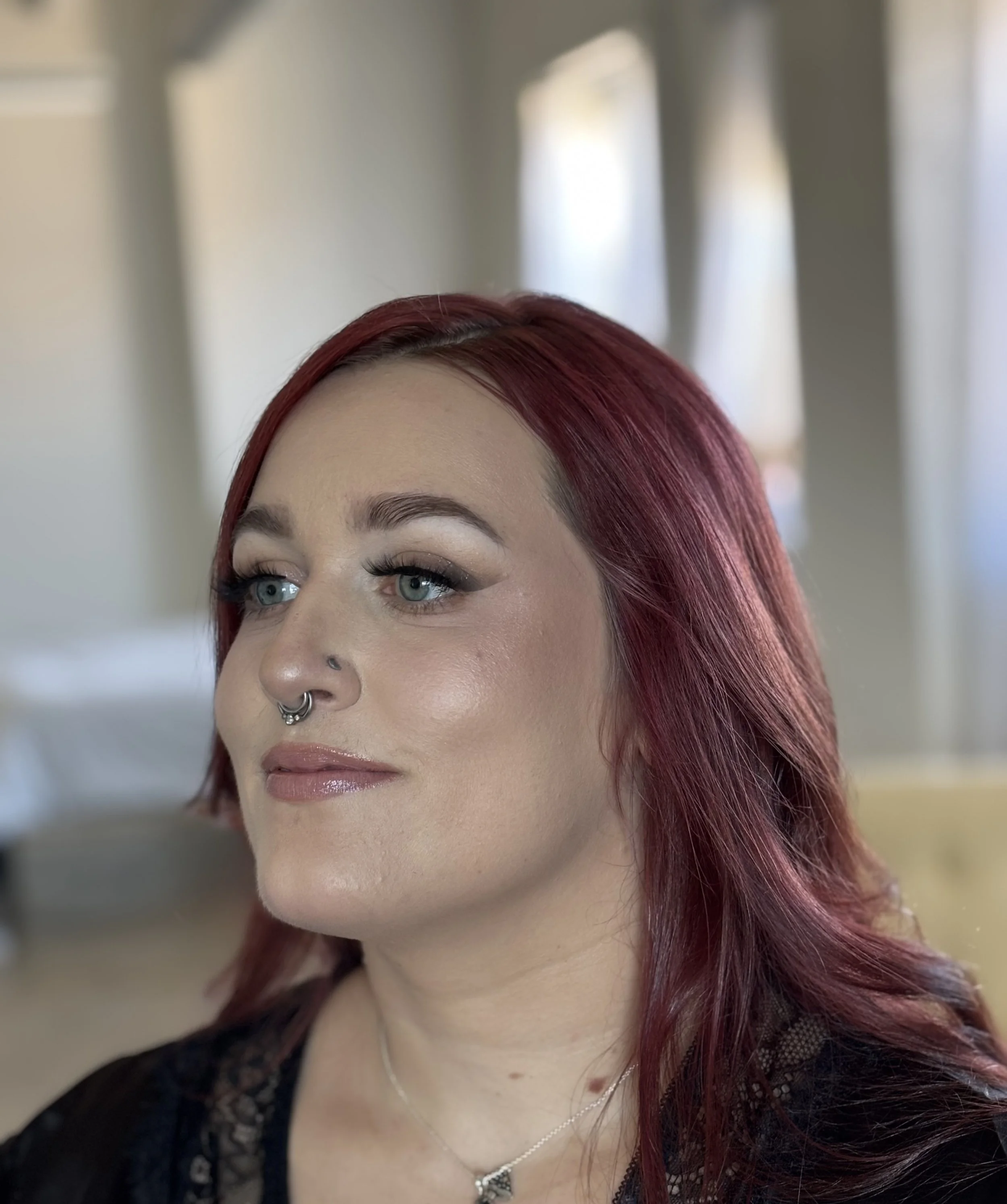 A close-up of a woman with red hair, green eyes, and a septum piercing, wearing a black lace top and a silver necklace with a heart-shaped pendant.