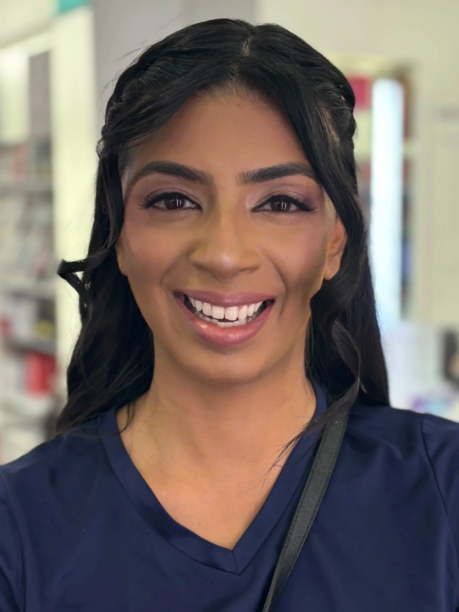 Close-up of a woman with long black hair, smiling, wearing dark blue scrubs, with a blurred background of a store or pharmacy setting.