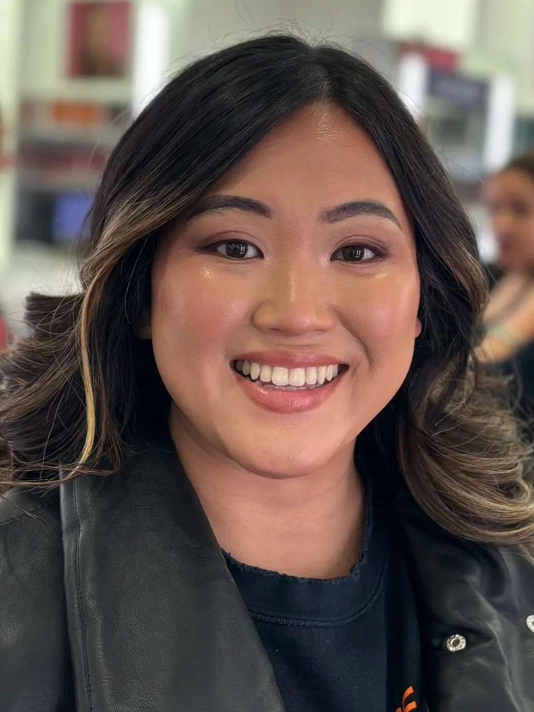 Smiling woman with black and brown hair, wearing a black jacket, indoors with blurred background.