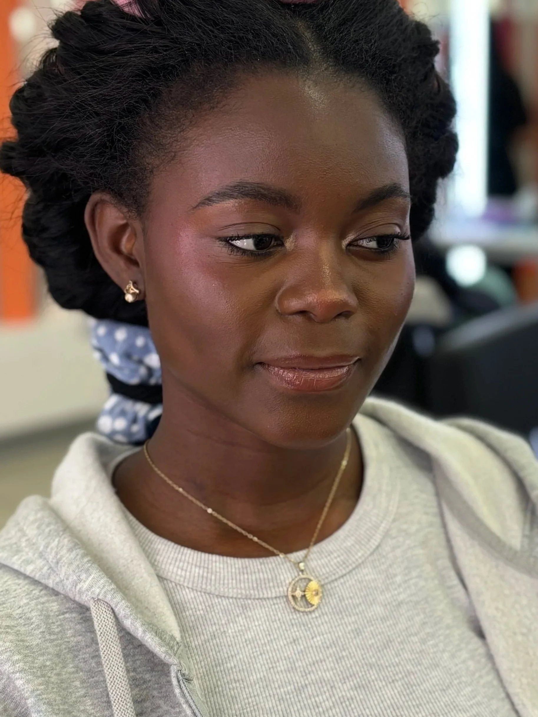 Close-up of a woman's face with dark skin and black hair styled in twists, wearing earrings and a layered necklace, in an indoor setting.