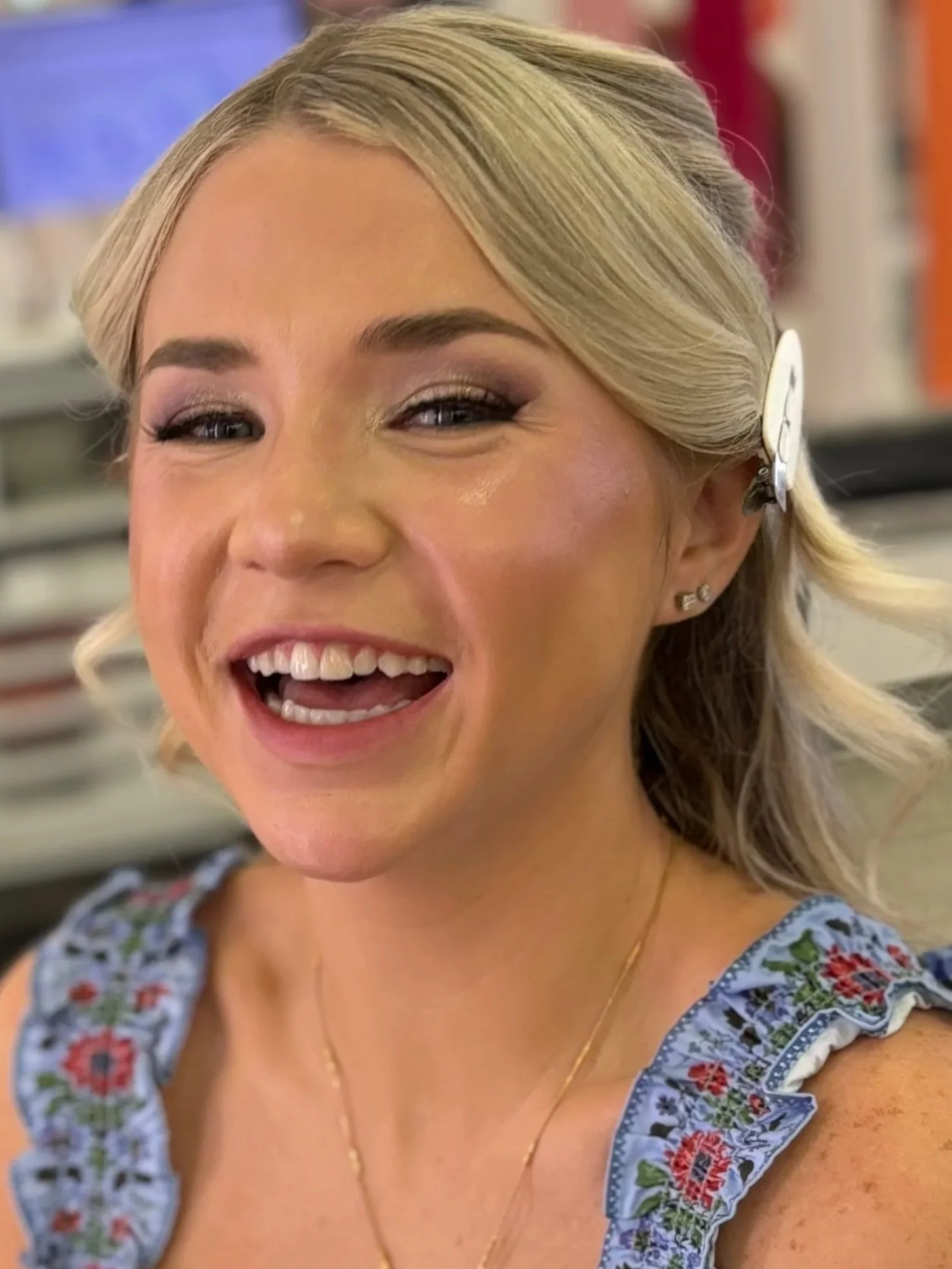 A smiling woman with blonde hair styled in loose waves, wearing a floral-patterned top with ruffled sleeves, small earrings, and a delicate necklace, standing in what appears to be a store.