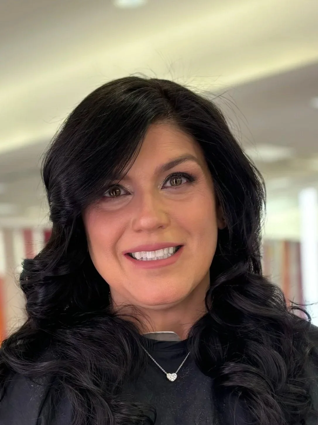 A woman with long dark curly hair, light skin, and makeup, smiling at the camera. She is wearing a black top, a silver necklace with a heart-shaped pendant, and is indoors with bookshelves in the background.