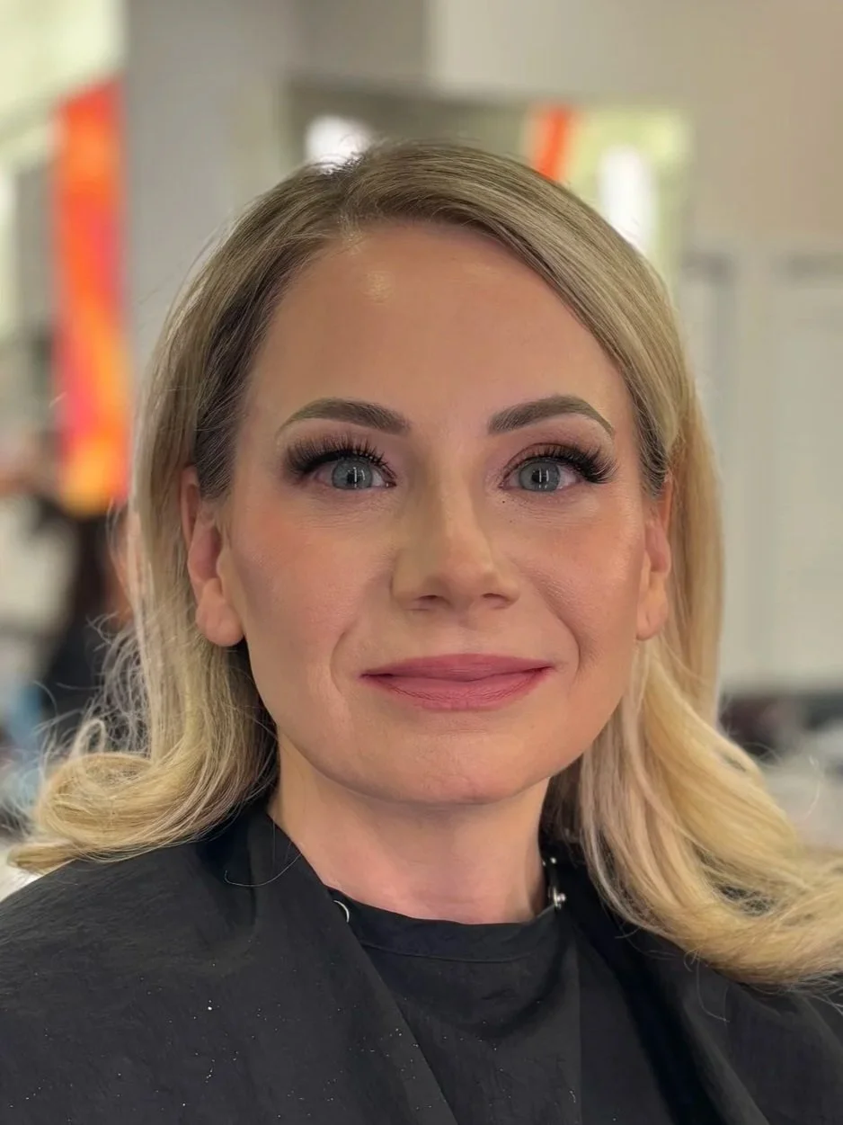 Close-up of a woman with blonde hair and blue eyes, smiling, in a salon or studio setting.
