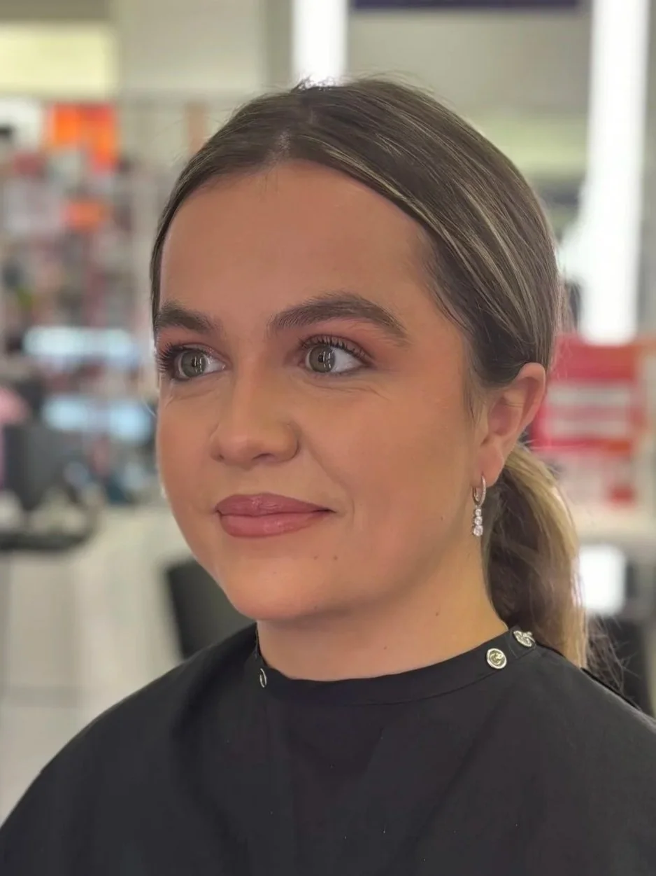Close-up of a woman with light brown hair, wearing makeup, earrings, and a black top with metal accents, inside a store or office environment.