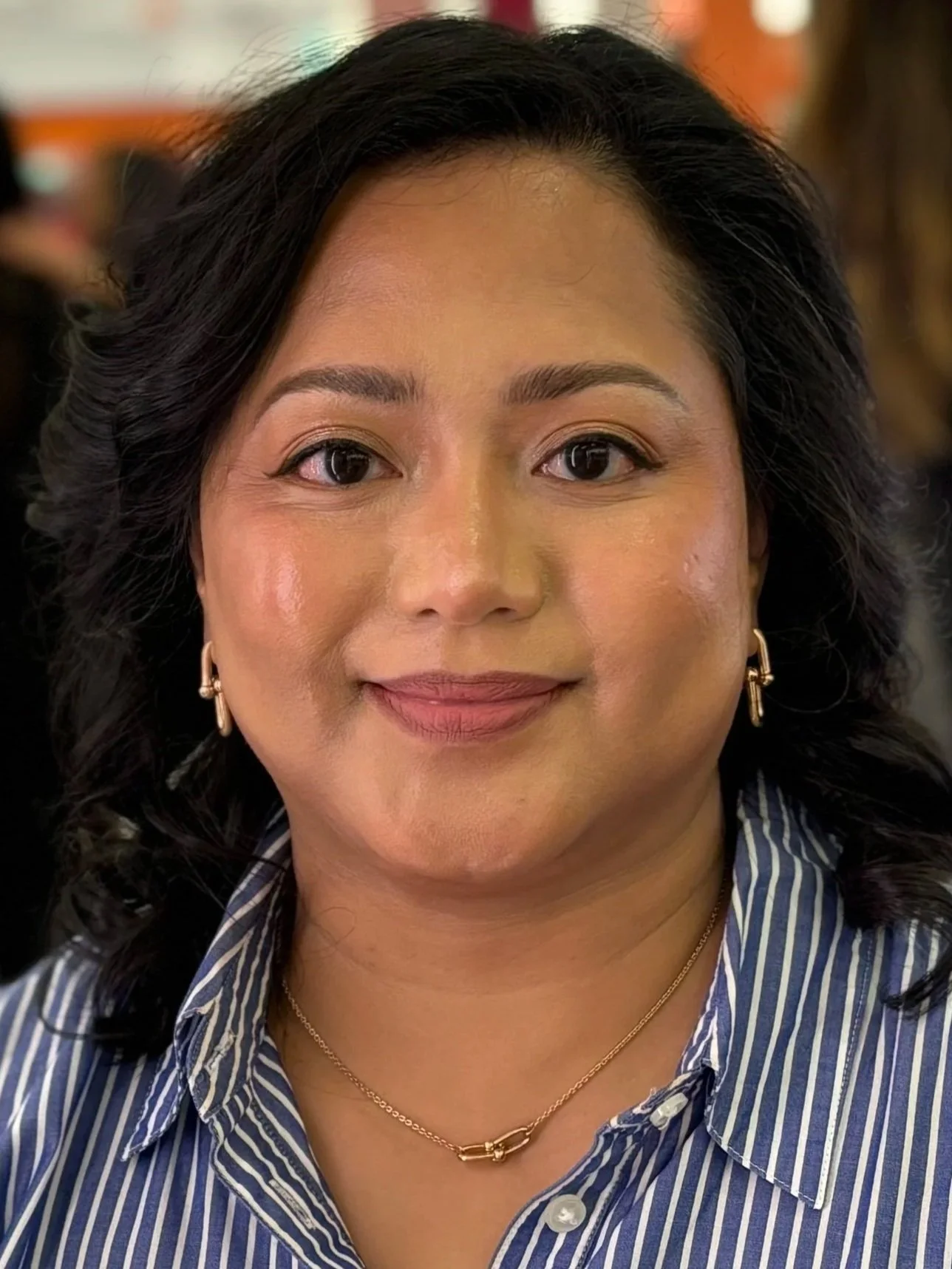 Close-up of a smiling woman wearing a striped shirt, gold earrings, and a gold necklace, with black curly hair, indoors with blurred background.