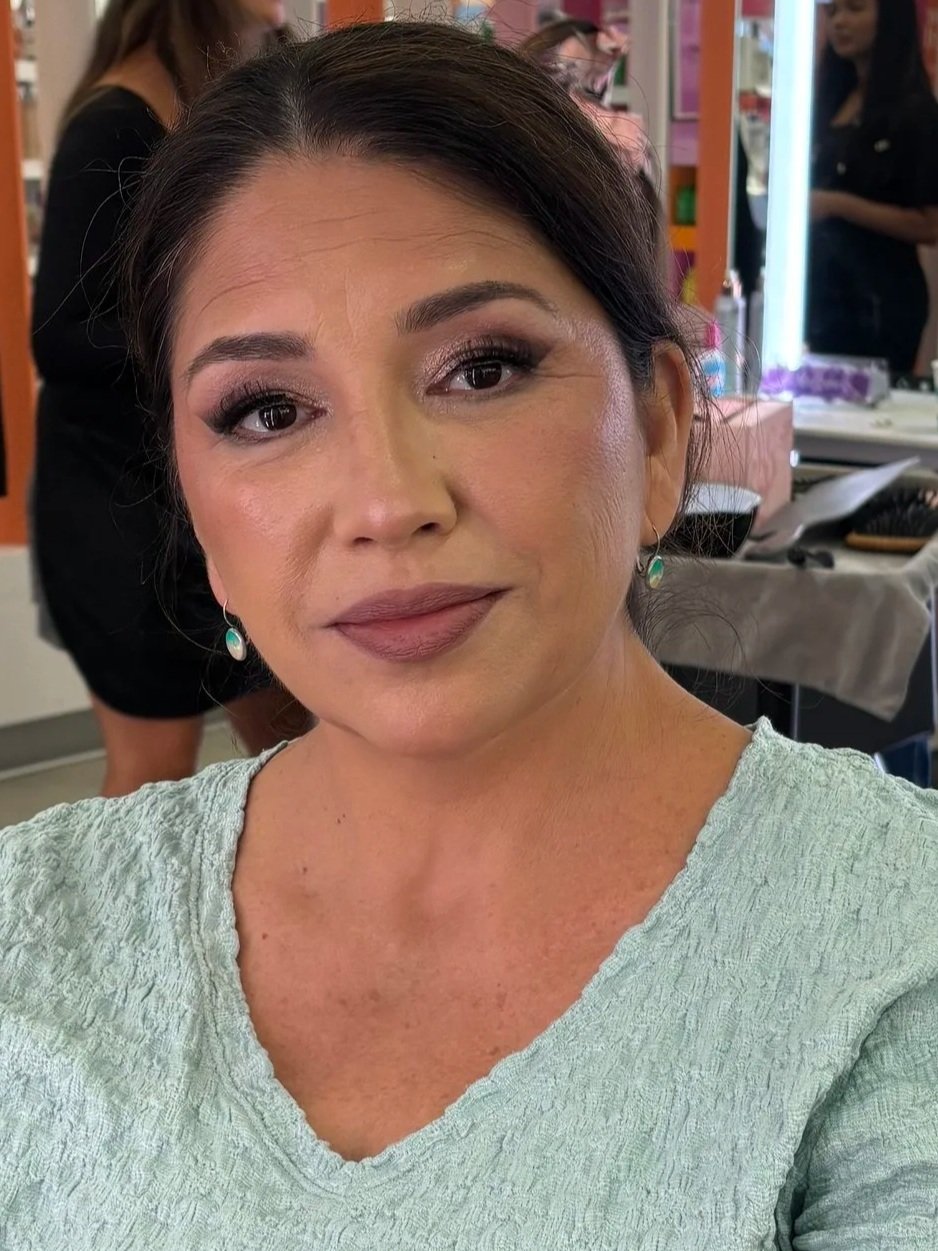 Close-up of a woman with makeup and earrings in a salon or makeup studio, with people and makeup tools in the background.