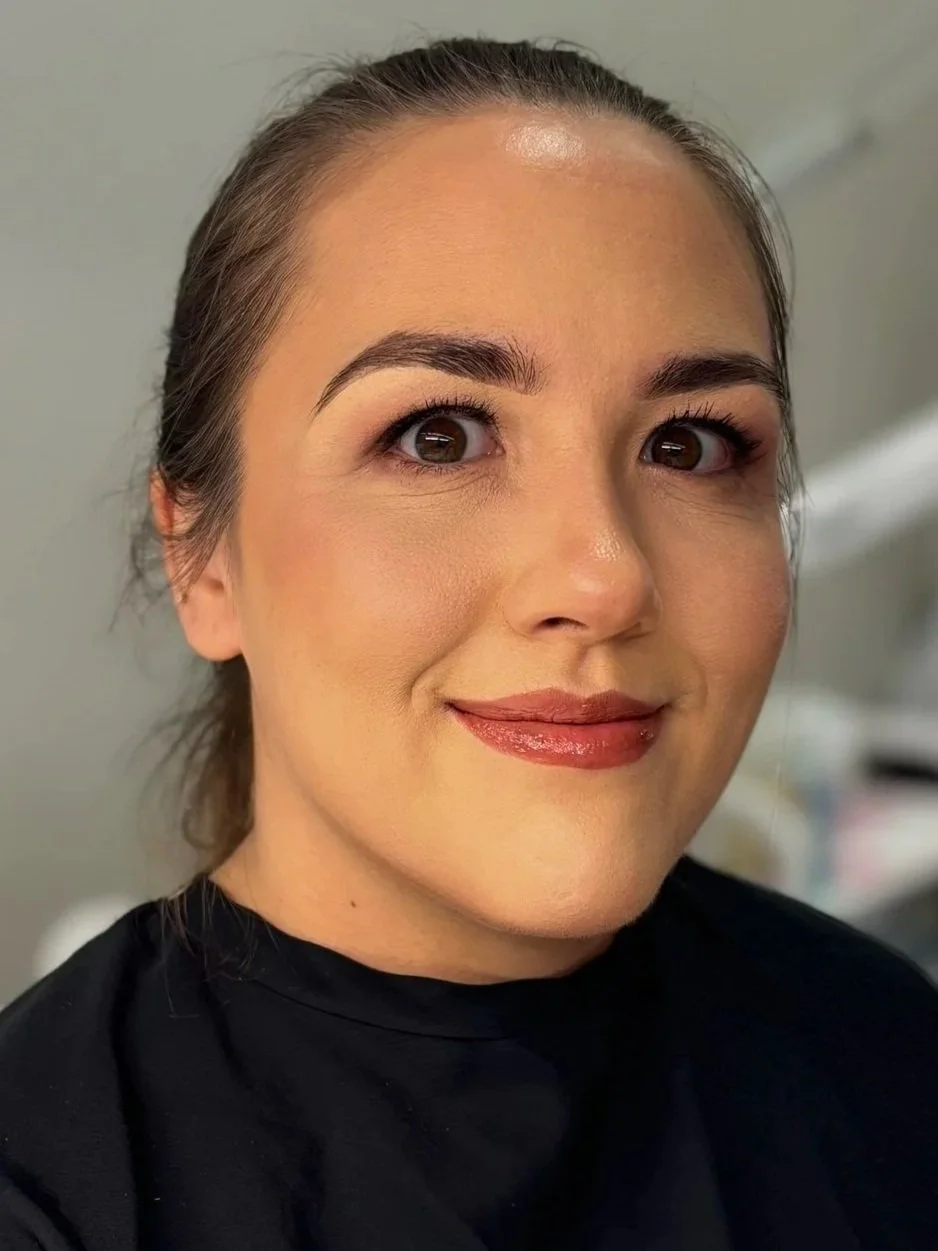 Close-up of a smiling woman with brown hair, wearing makeup, and a black top, indoors.