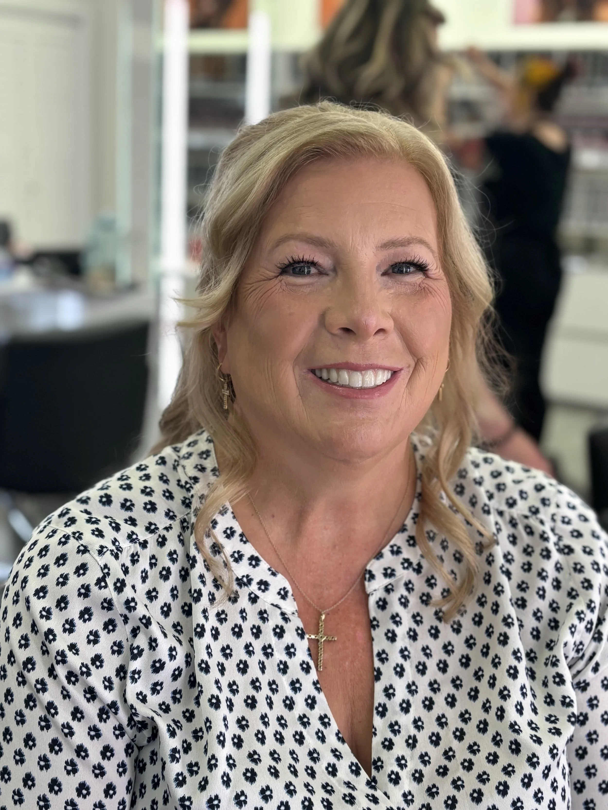Close-up of a smiling woman with blonde hair, blue eyes, wearing a white blouse with navy floral pattern, a cross necklace, and gold earrings, in an indoor setting with blurred background.