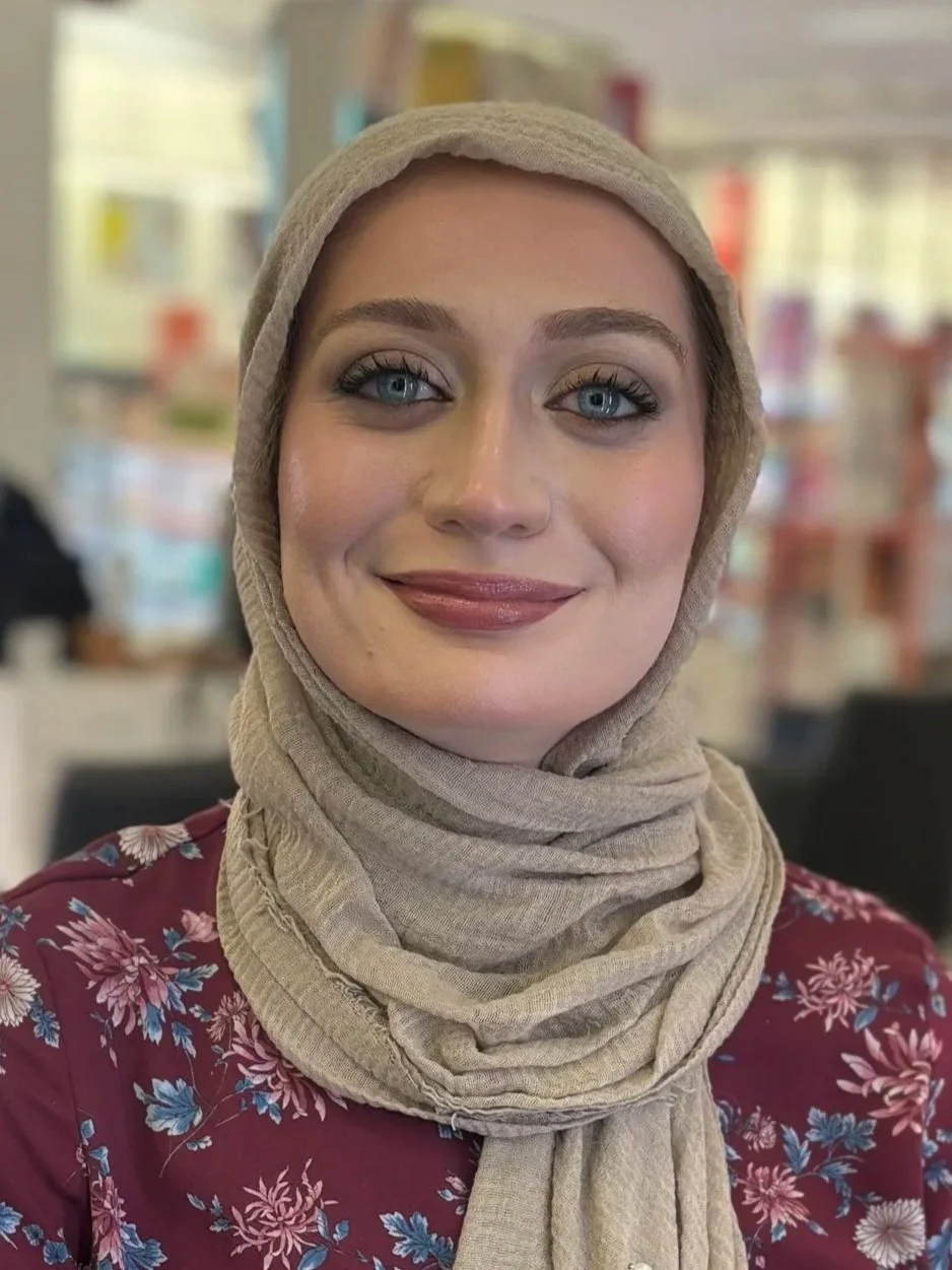 Close-up of a woman with blue eyes and light makeup wearing a beige headscarf and a floral red top, smiling in a bright indoor setting.