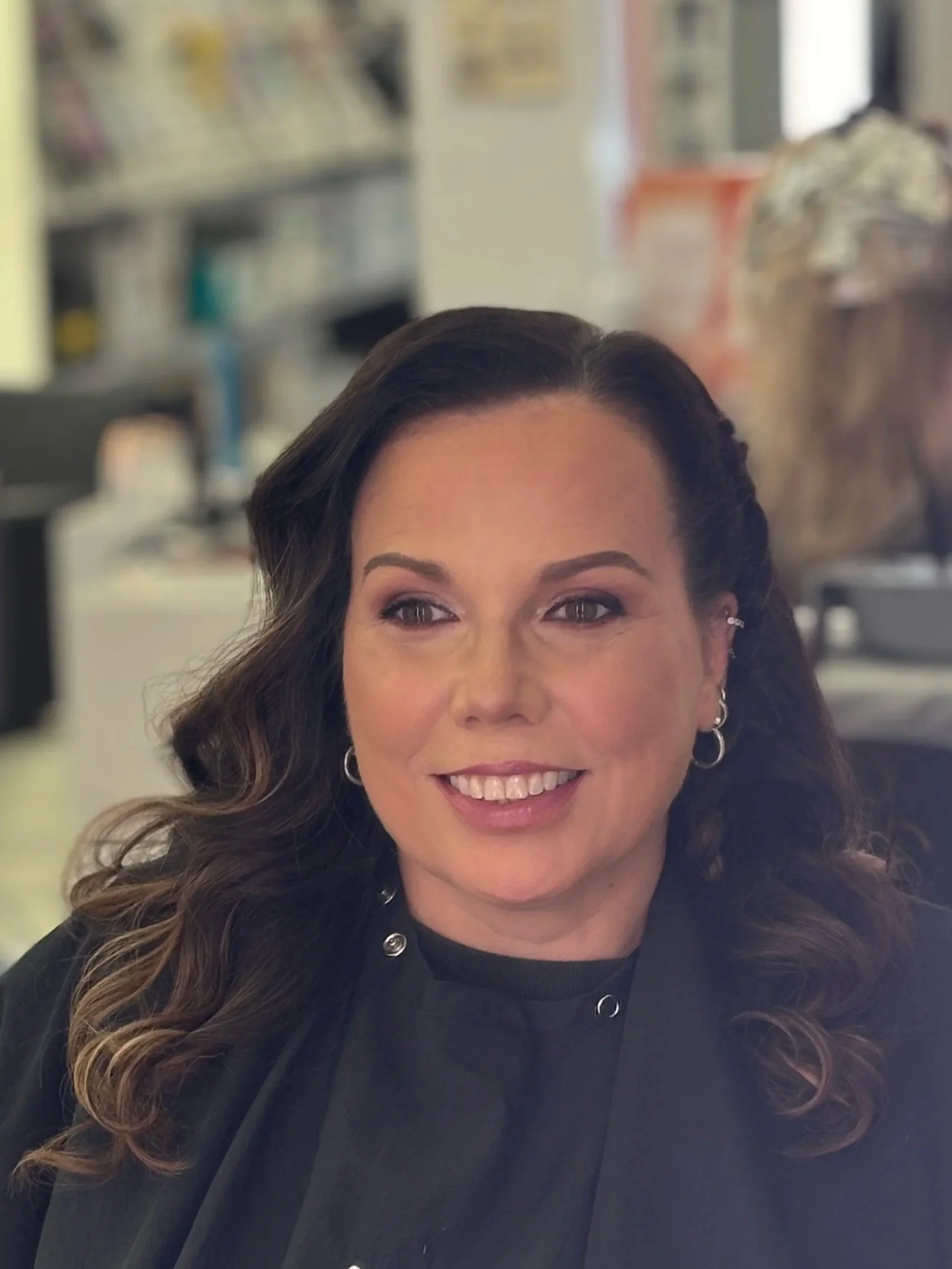 Close-up of a woman’s face, smiling, with long curly hair, wearing earrings and a black top, in an indoor setting with blurred background.