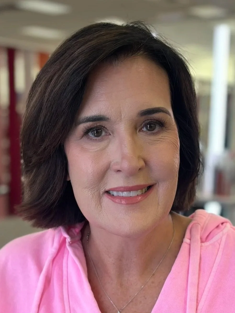 Close-up of a woman with dark brown hair, wearing a pink top, smiling in an indoor setting.