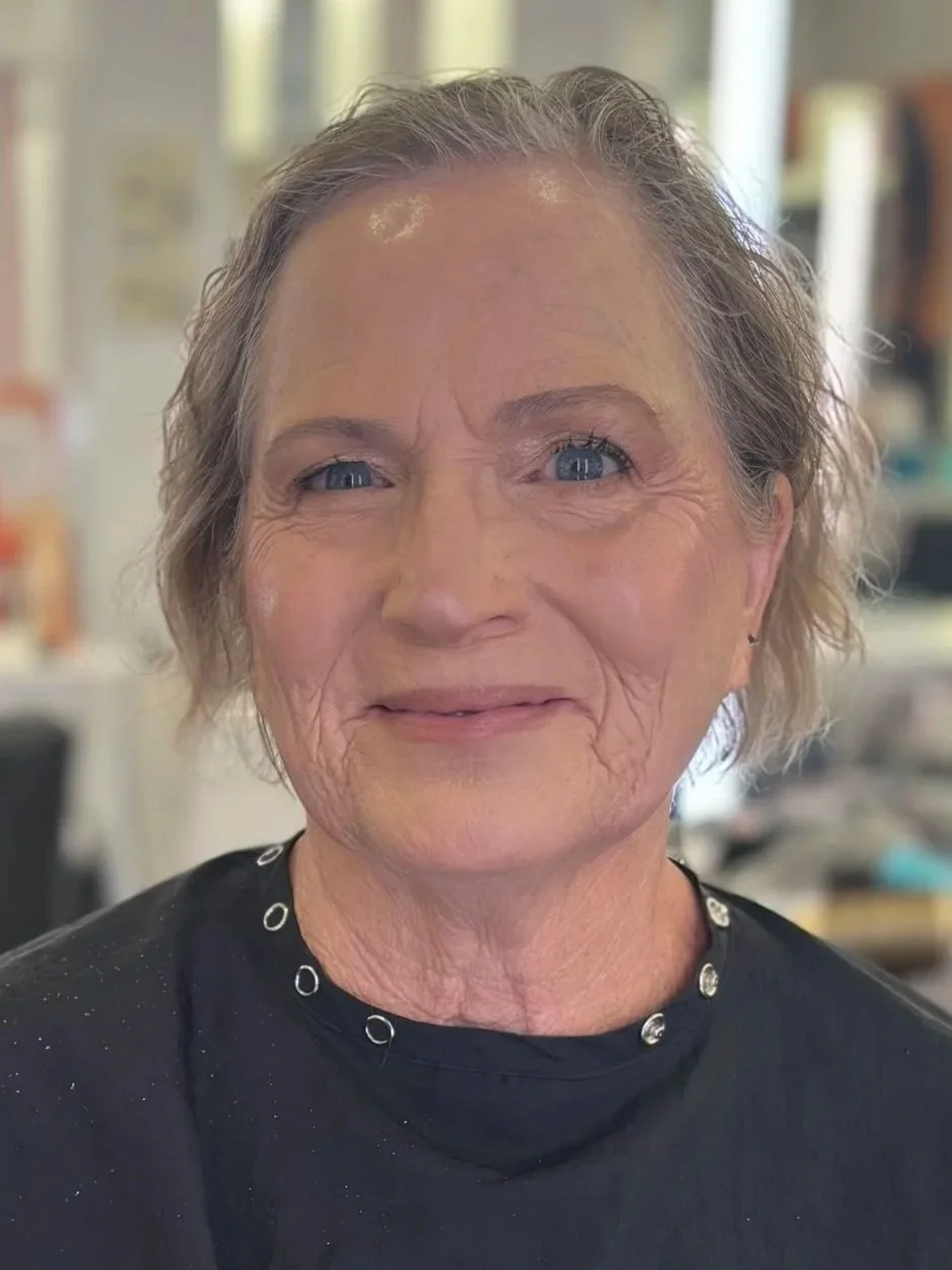 Close-up of an elderly woman with short gray hair, blue eyes, and visible wrinkles, smiling in an indoor setting.