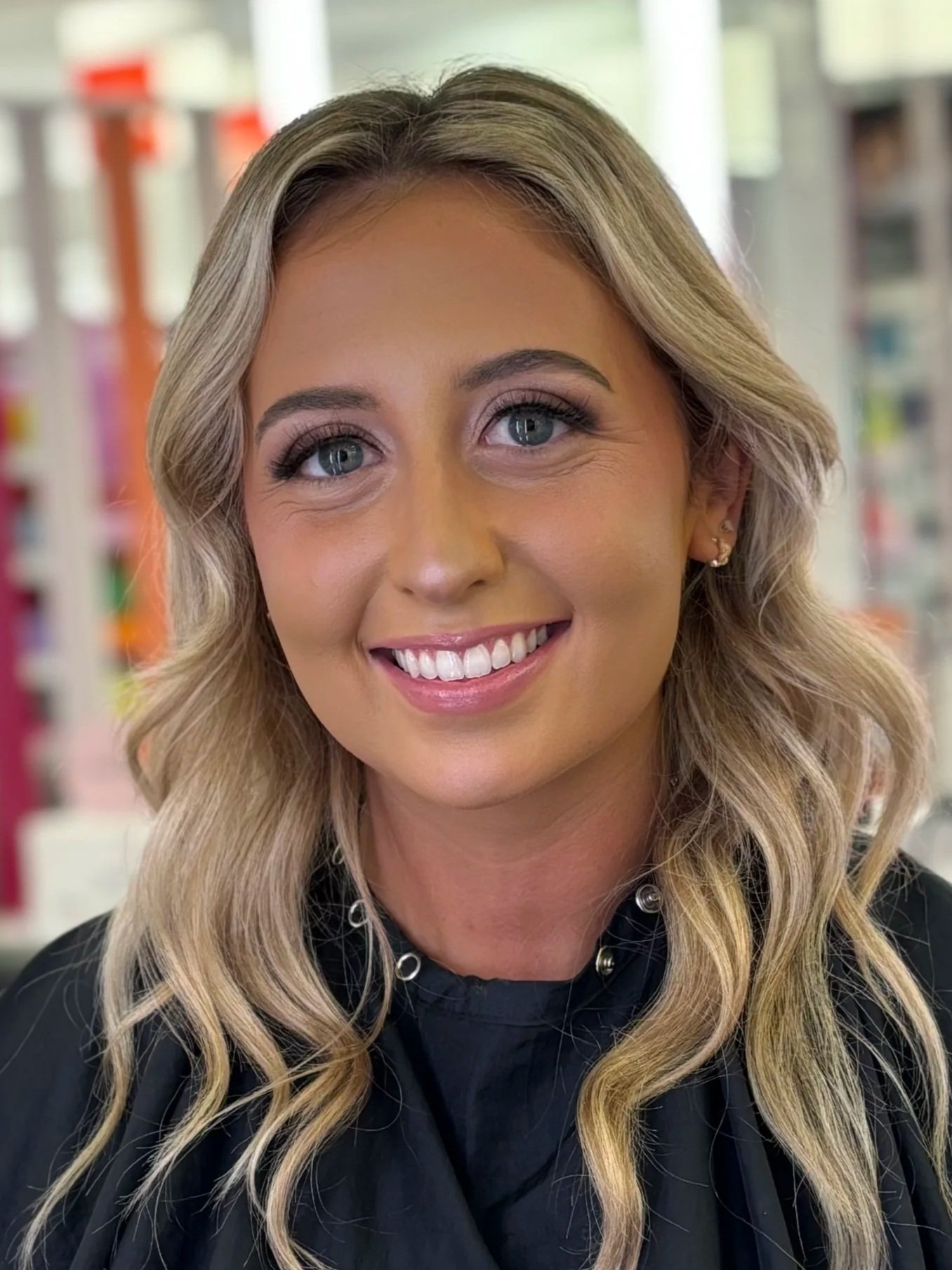 A woman with long, wavy blonde hair, blue eyes, and a bright smile, wearing makeup and a black top with pearl necklaces, in a brightly lit environment with shelves and store items in the background.