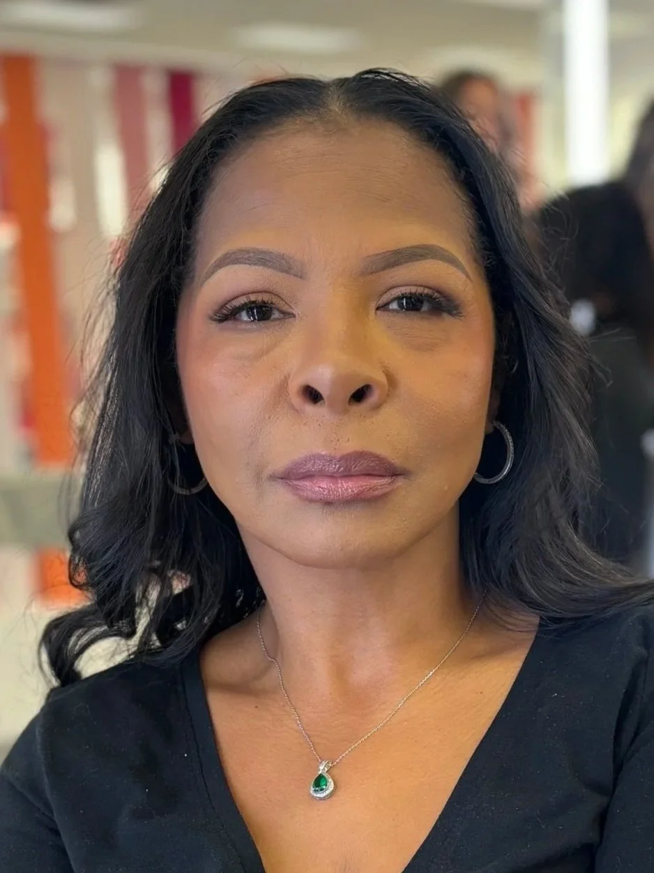 A woman with black wavy hair, wearing hoop earrings and a silver necklace with a green pendant, taking a selfie indoors.