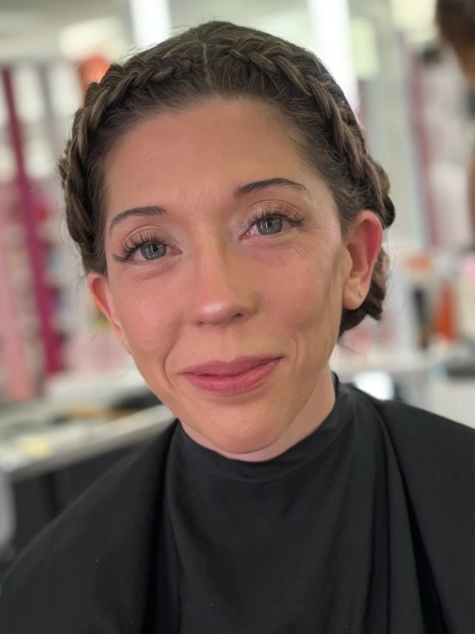 Close-up of a woman with braided hair, blue eyes, and wearing a black top, smiling slightly, in what appears to be a salon or makeup area.