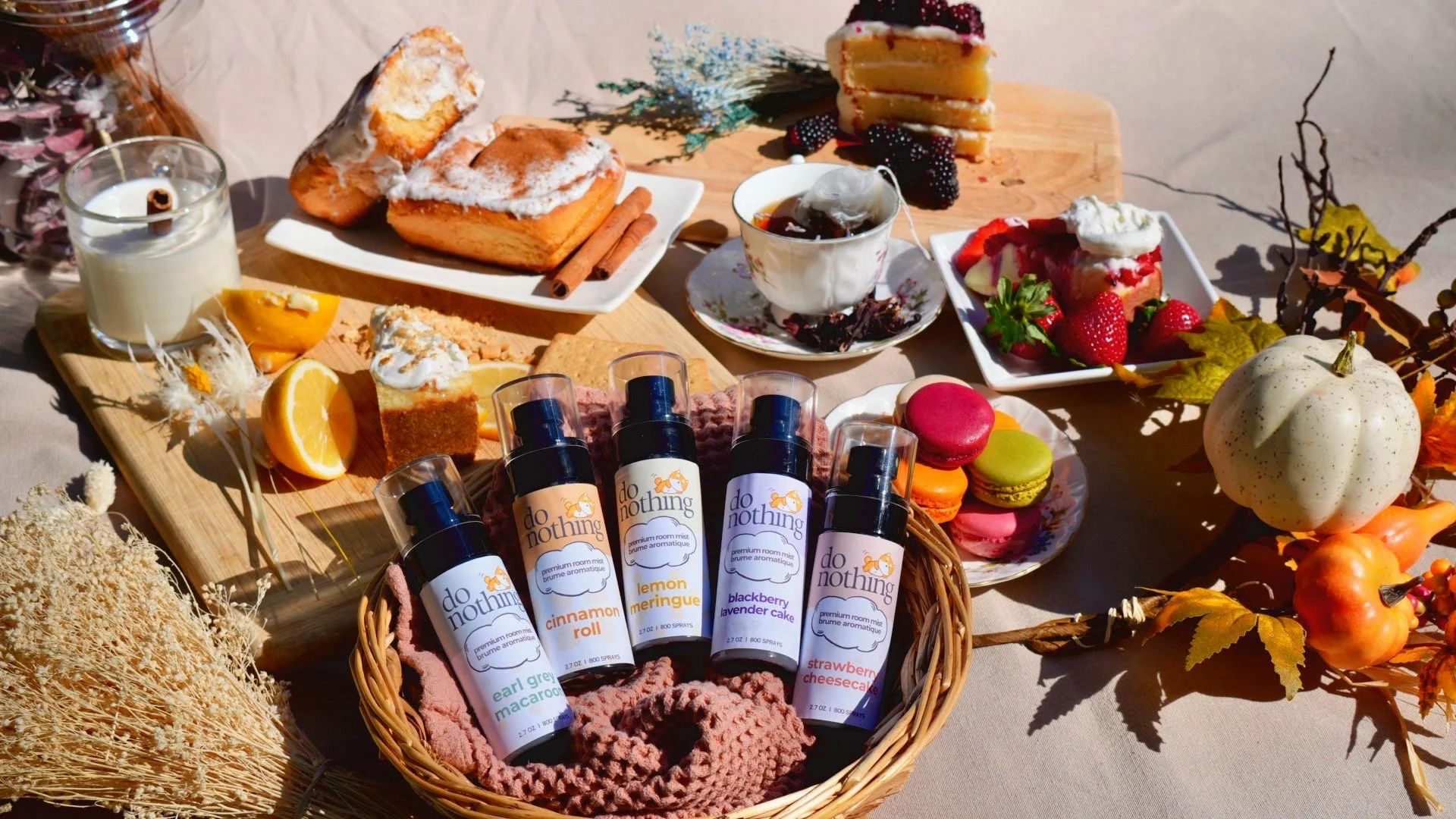 A table set with desserts, drinks, and decorative fall elements. Includes cakes, macarons, a lemon meringue pie, strawberries, orange slices, and a white decorative pumpkin, with scented room mist bottles in the foreground.