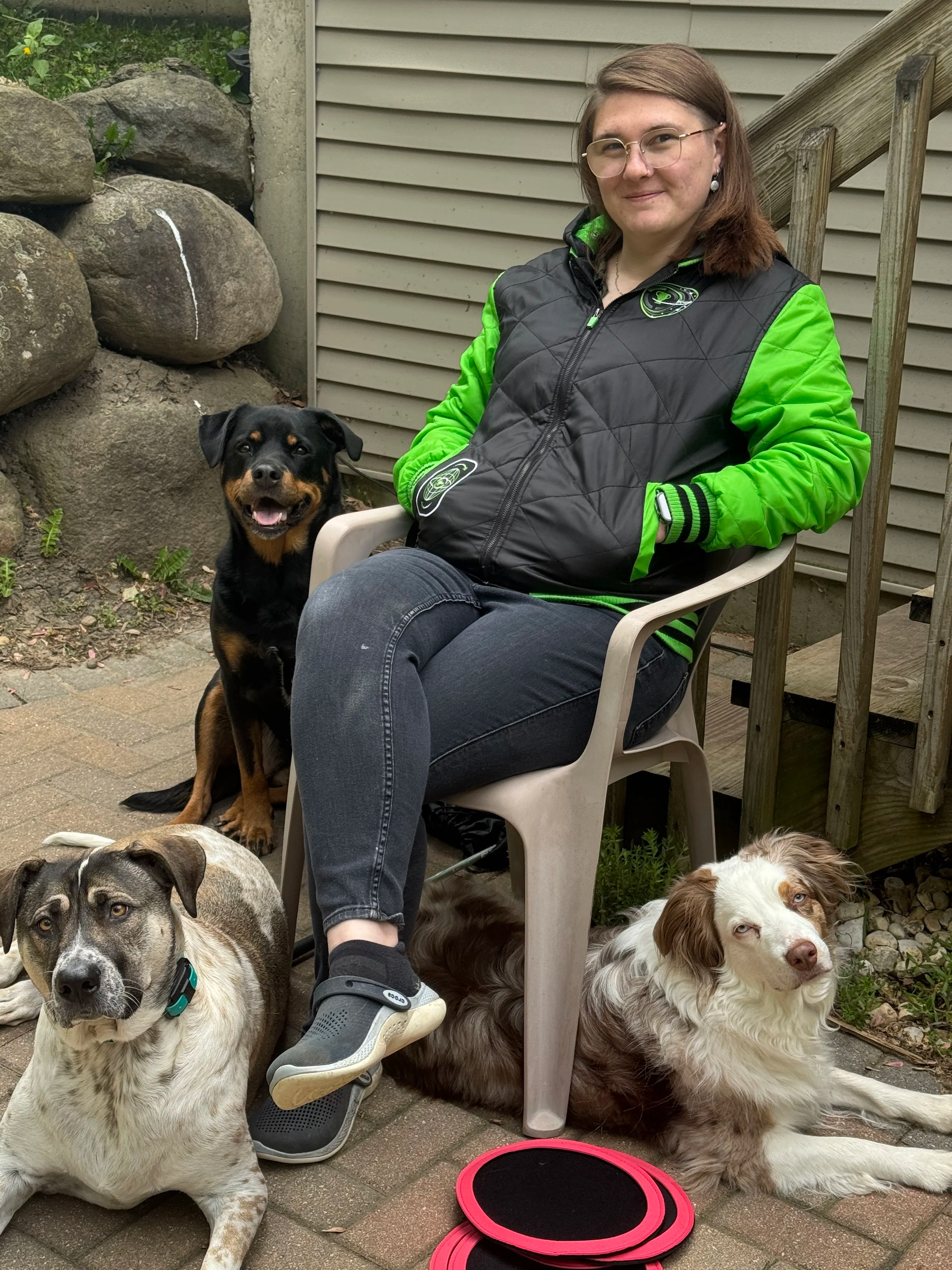 A woman sitting on a plastic chair outdoors with four dogs around her, in front of a house with a wooden staircase and rocks wall, during daytime.