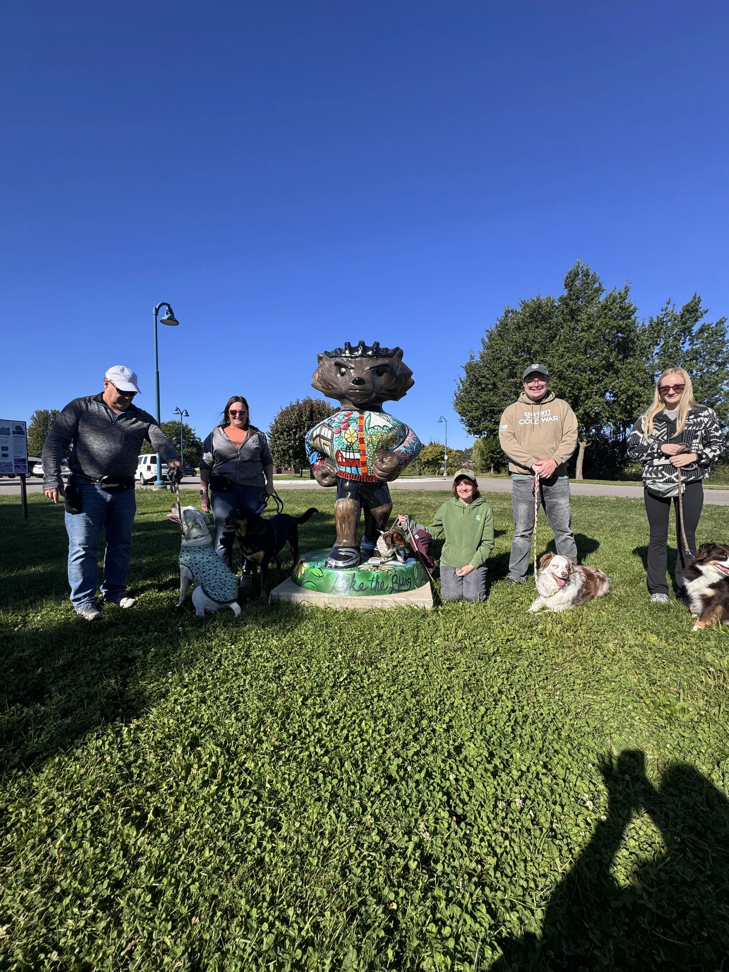 Group of five people and four dogs standing around a colorful statue of a hedgehog on a grassy park under clear blue sky.