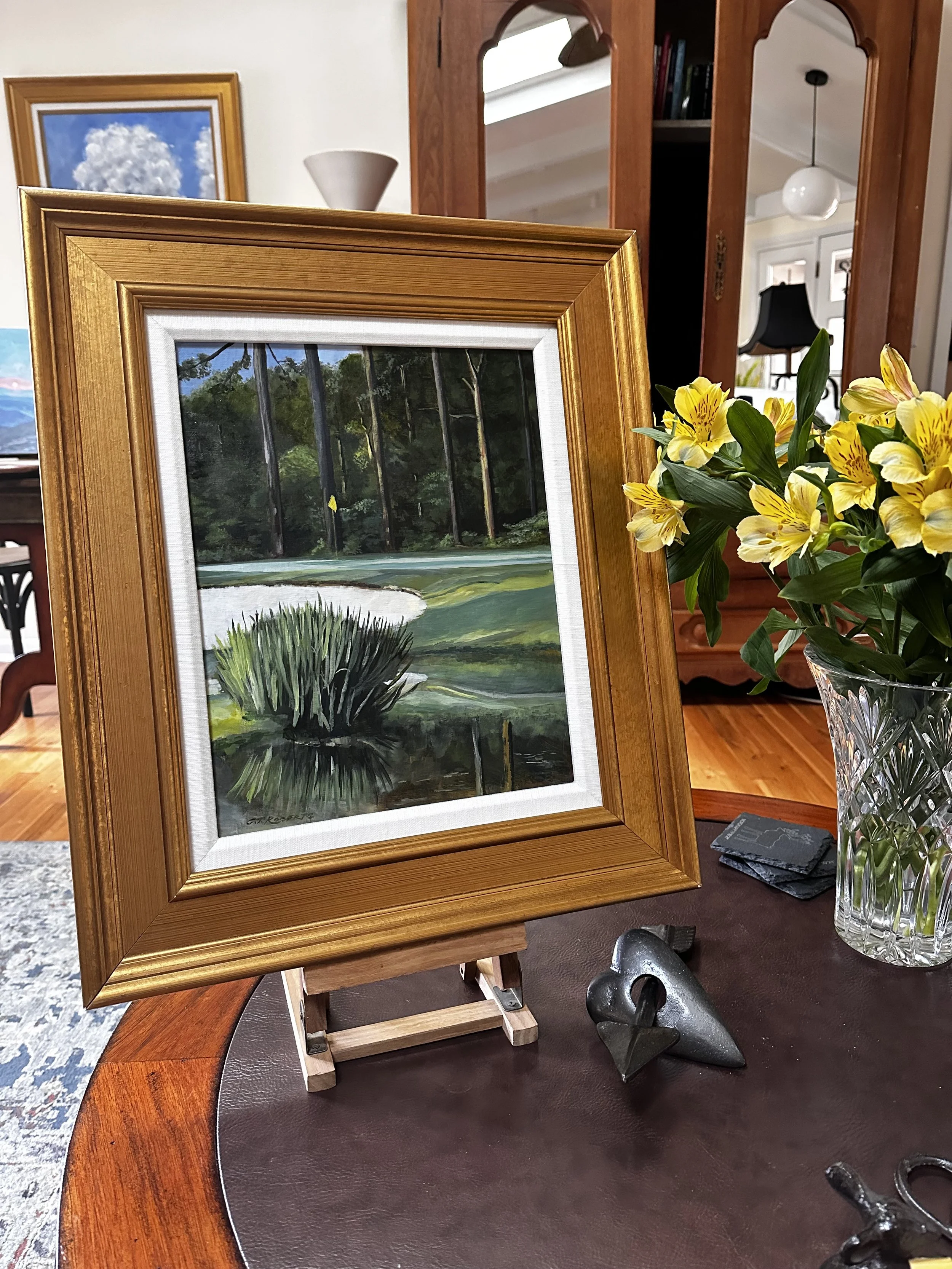 A framed landscape painting of a golf course scene with trees, a pond, and a sand trap, placed on a small easel on a wooden table. There is a vase with yellow flowers to the right of the painting and a small heart-shaped sculpture on the table.