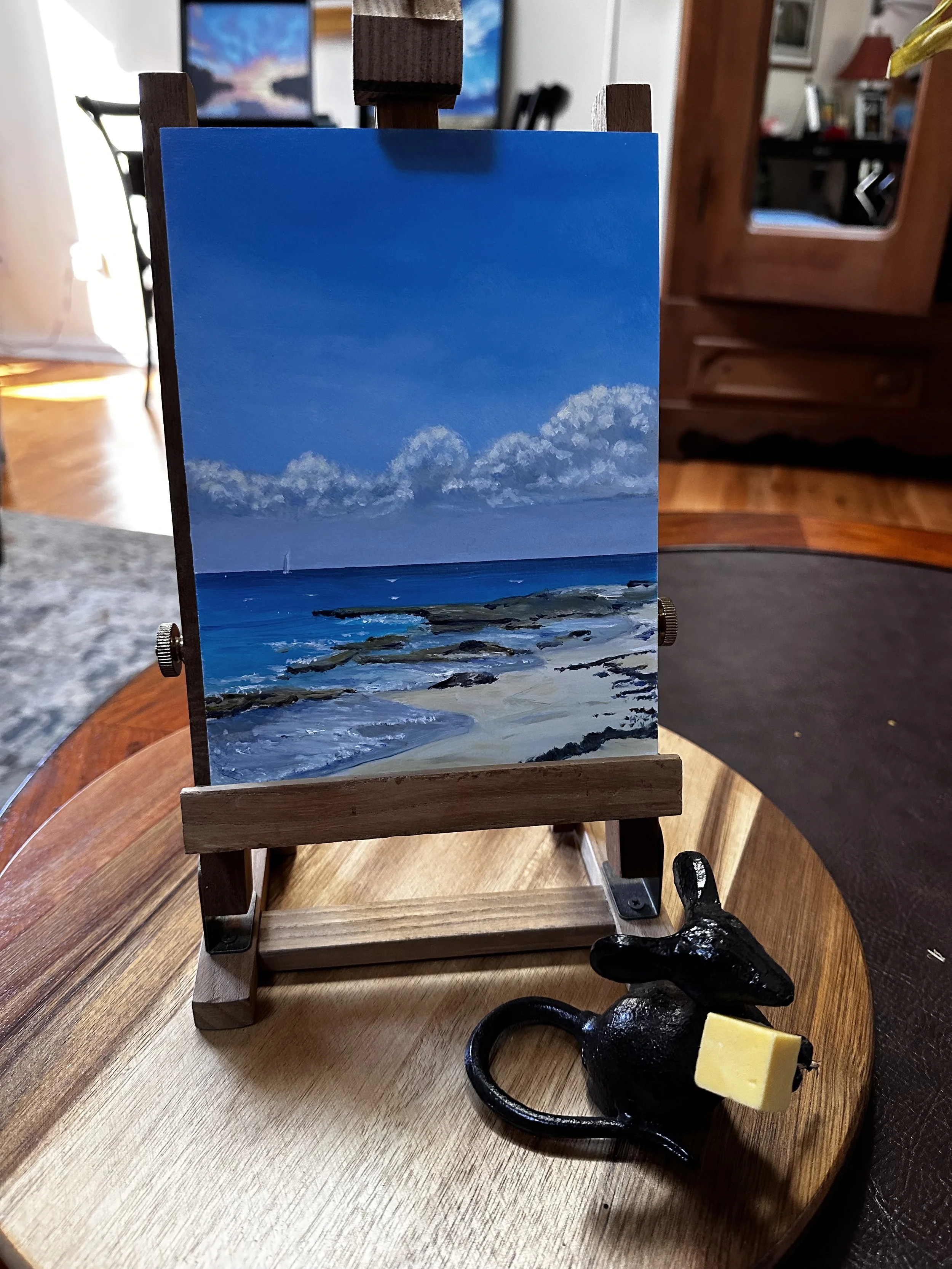 Miniature easel holding a small landscape painting of a beach with rocks, blue water, and a sky filled with clouds, positioned on a wooden table alongside a black rabbit figurine with a yellow piece attached to its ear.