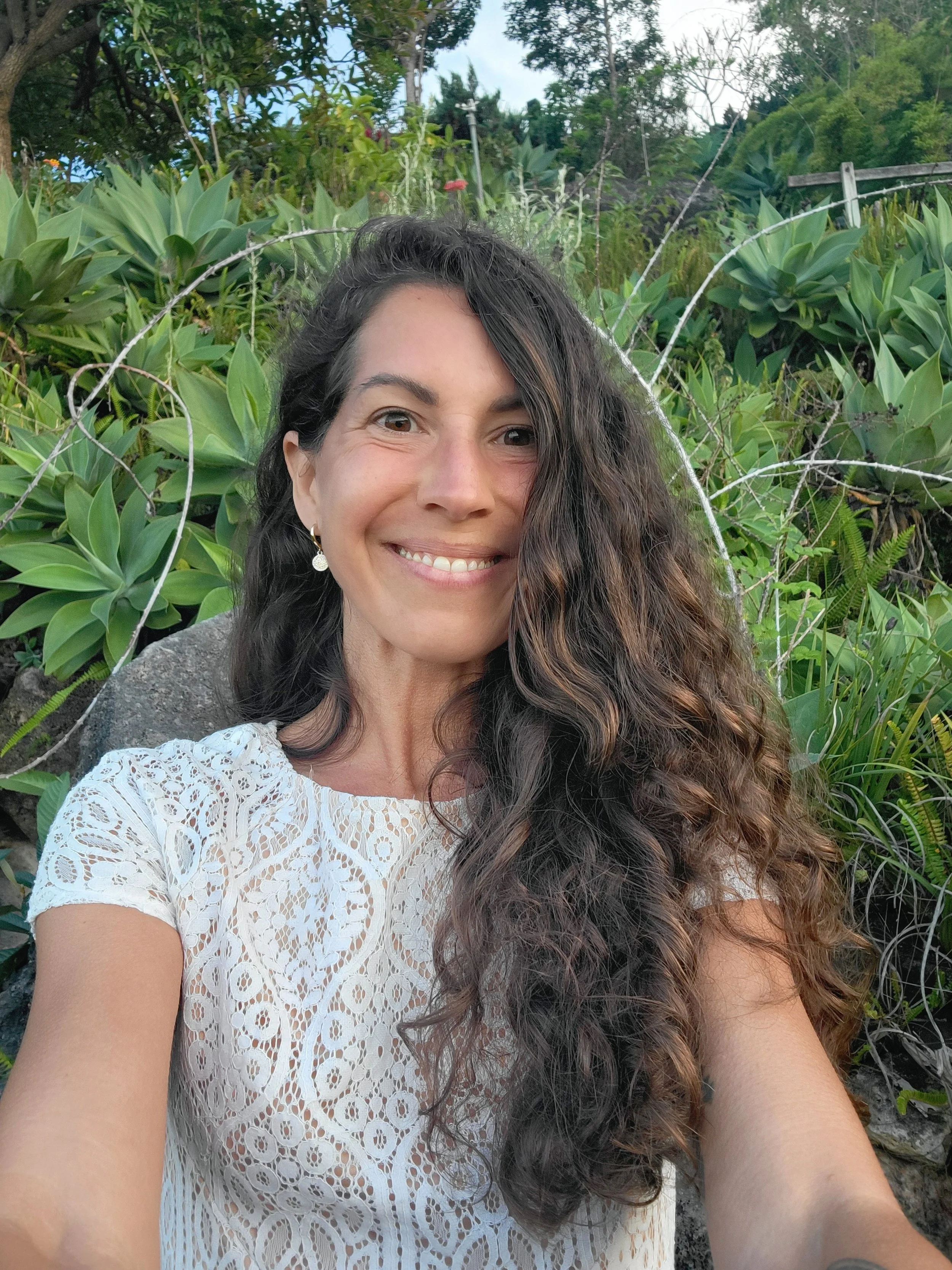 Une femme souriante avec de long cheveux bouclés, portant un haut blanc en dentelle, posant devant une végétation luxuriante.