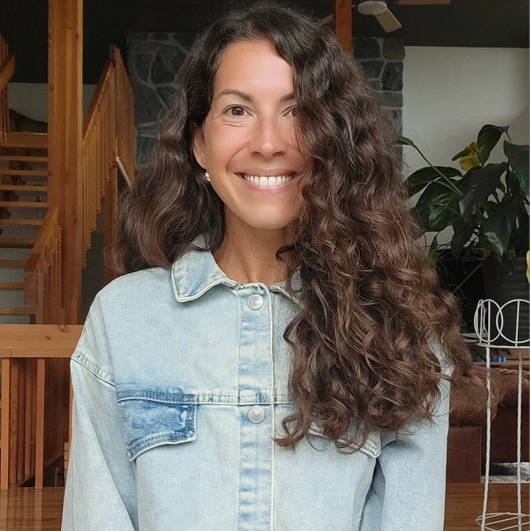 Femme souriante à la maison, portant une veste en jean avec un intérieur en bois et une plante verte en arrière-plan.