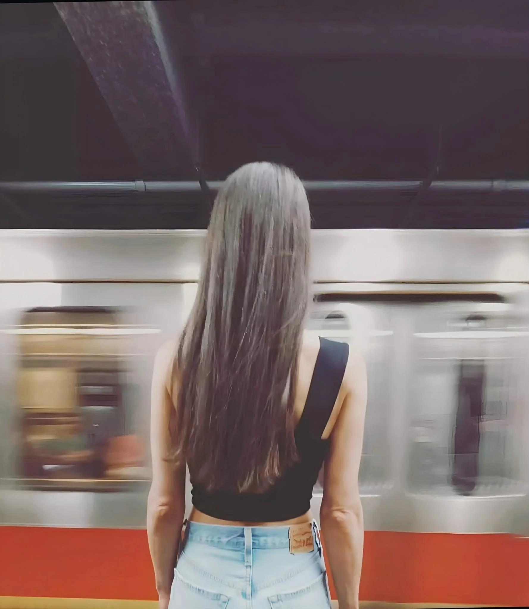 Une femme avec des cheveux longs et ondulés se tient devant un métro en mouvement dans une station.