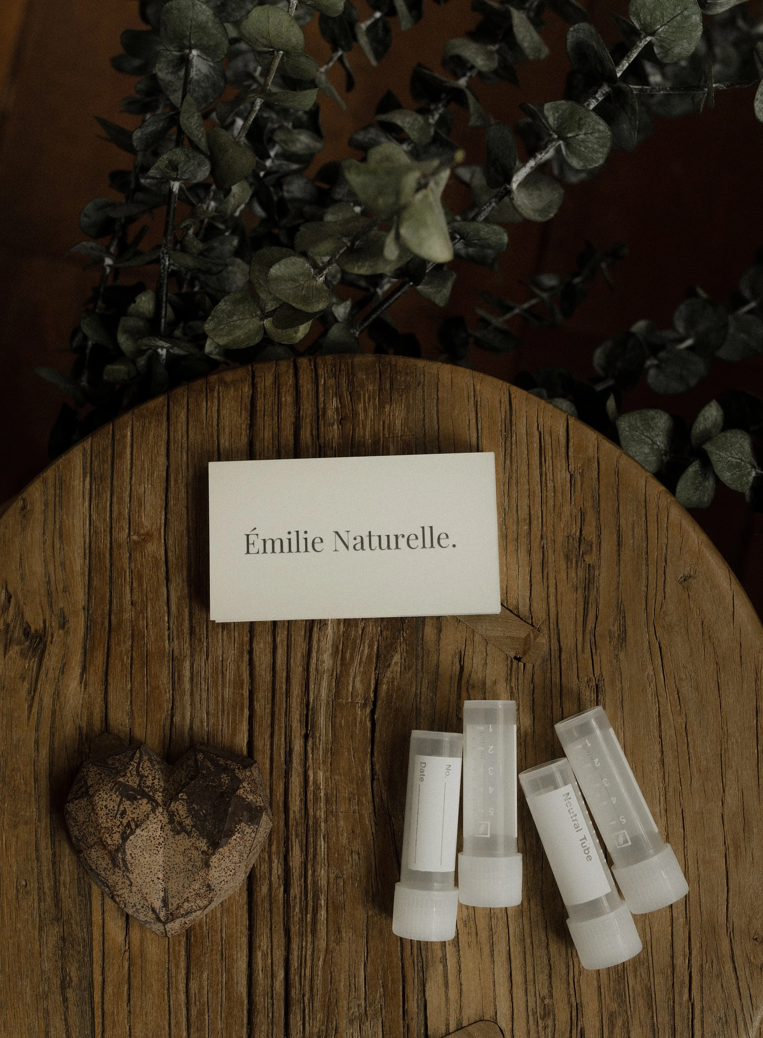 Une petite boîte en forme de cœur en pierre, trois tubes de produit avec étiquettes, une grande feuille avec un label 'Émilie Naturelle', et un feuillage vert en arrière-plan.