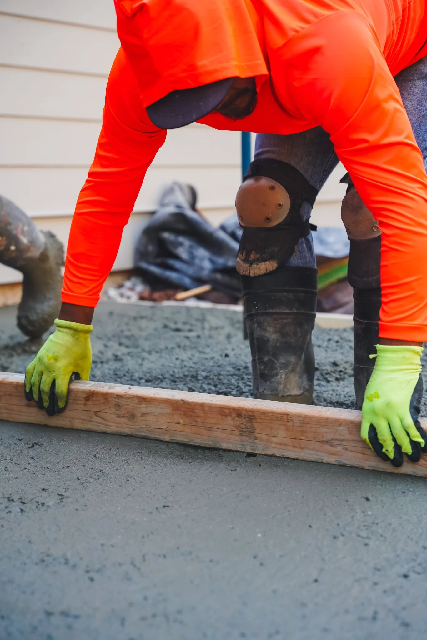 A construction worker wearing a black cap, orange long-sleeve shirt, black knee pads, and yellow gloves, is kneeling and leveling wet concrete with a wooden board on a construction site.
